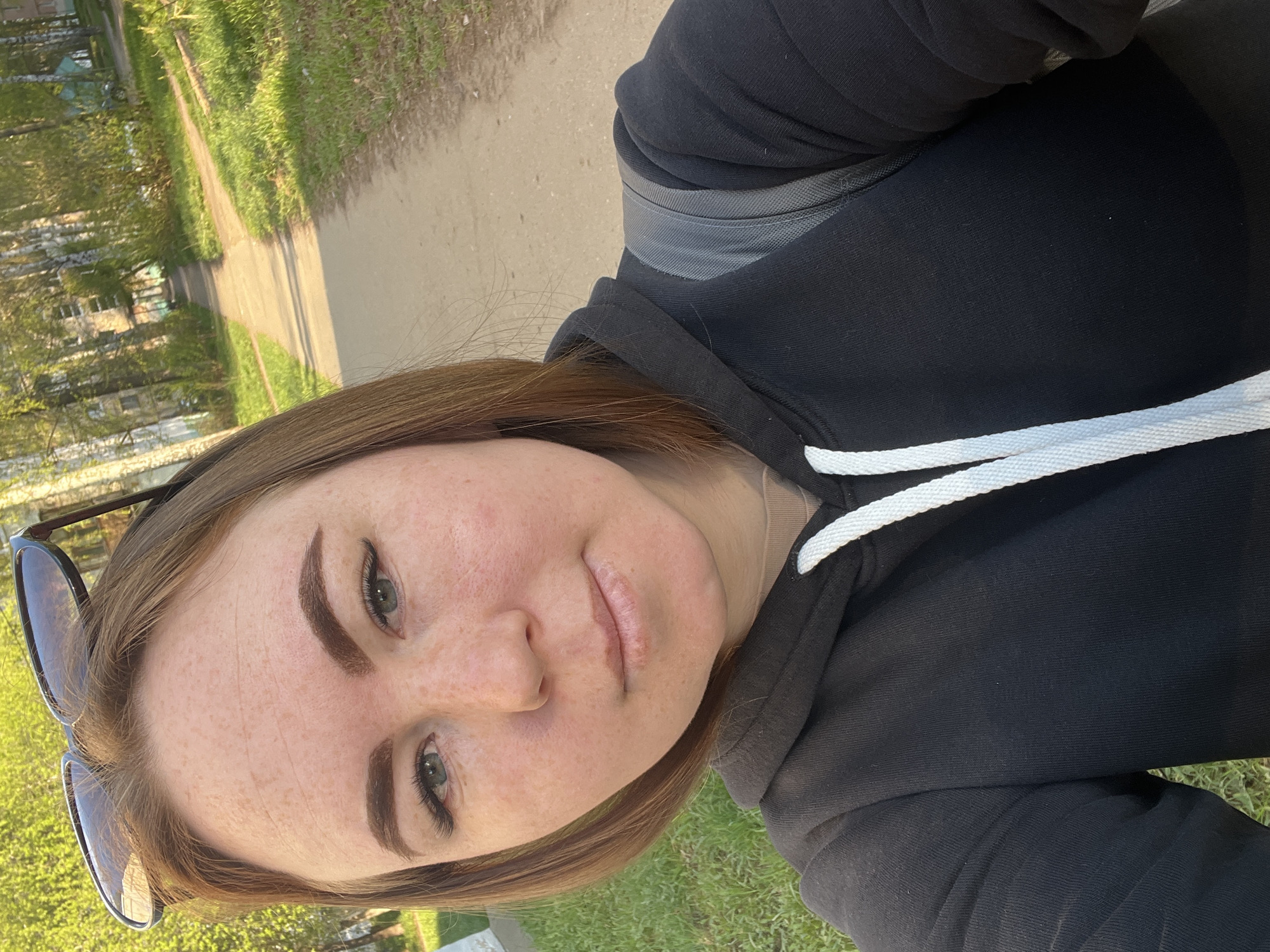A young woman is taking a selfie outdoors. She is wearing a black hooded sweatshirt with white drawstrings. Sunglasses are perched on her head. The background shows a paved path bordered by green grass and trees. The location is likely in the city of Chaikovsky, Russia, based on the user's prompt.