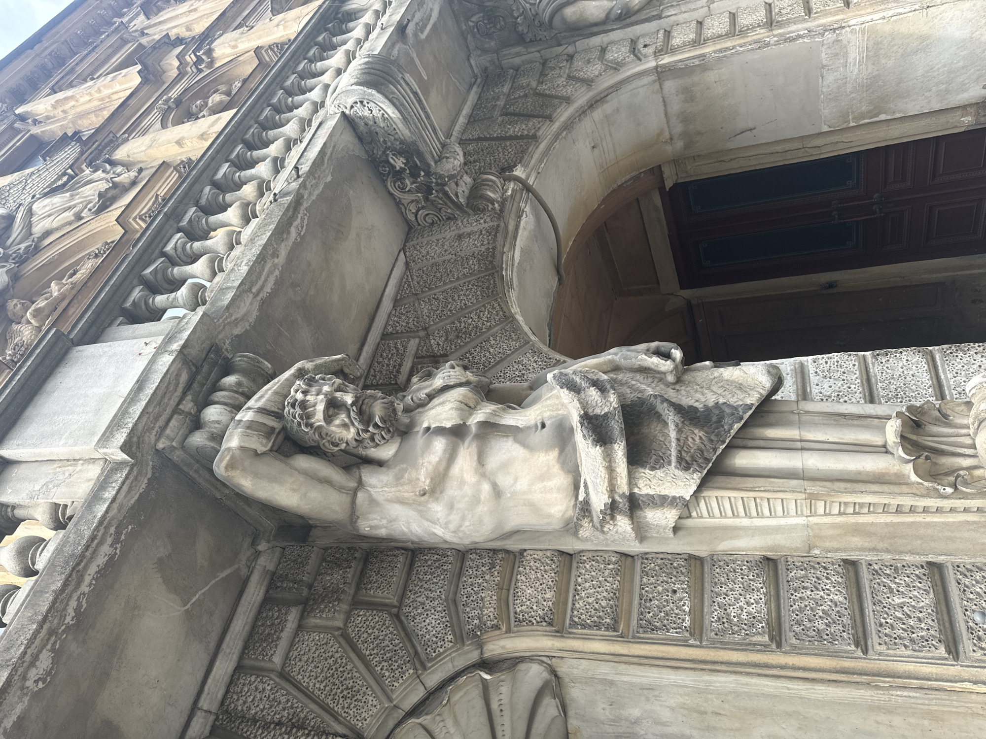The image captures a low-angle view of an ornate architectural facade, showcasing a detailed stone sculpture and elaborate decorative elements. The primary subject is a large, reclining male figure sculpted in stone, positioned horizontally above an archway. The figure is partially draped with fabric, revealing a muscular torso and an arm raised to its head, suggesting a pensive or weary pose. The intricate carving of his hair and features is visible.
Surrounding the sculpture and extending upwards is a richly decorated building facade, likely made of light-colored stone or marble. The architecture features classical elements, including a balustrade with multiple carved balusters, and various levels of decorative molding and cornices. The archway below the reclining figure is deeply recessed, with an elaborate, almost stippled or textured pattern carved into its spandrels and soffit. Inside the archway, a dark, presumably wooden door or entrance is visible, characterized by rectangular panels and some subtle detailing.
The perspective is from below, looking up at the building, emphasizing its grandeur and height. The lighting appears soft and even, suggesting an overcast day or indirect light, which highlights the textures and depth of the carvings without harsh shadows. The stone shows signs of age and weathering, contributing to the historical character of the structure. No visible text is present in the image. The scene suggests a historically significant building in an urban setting.