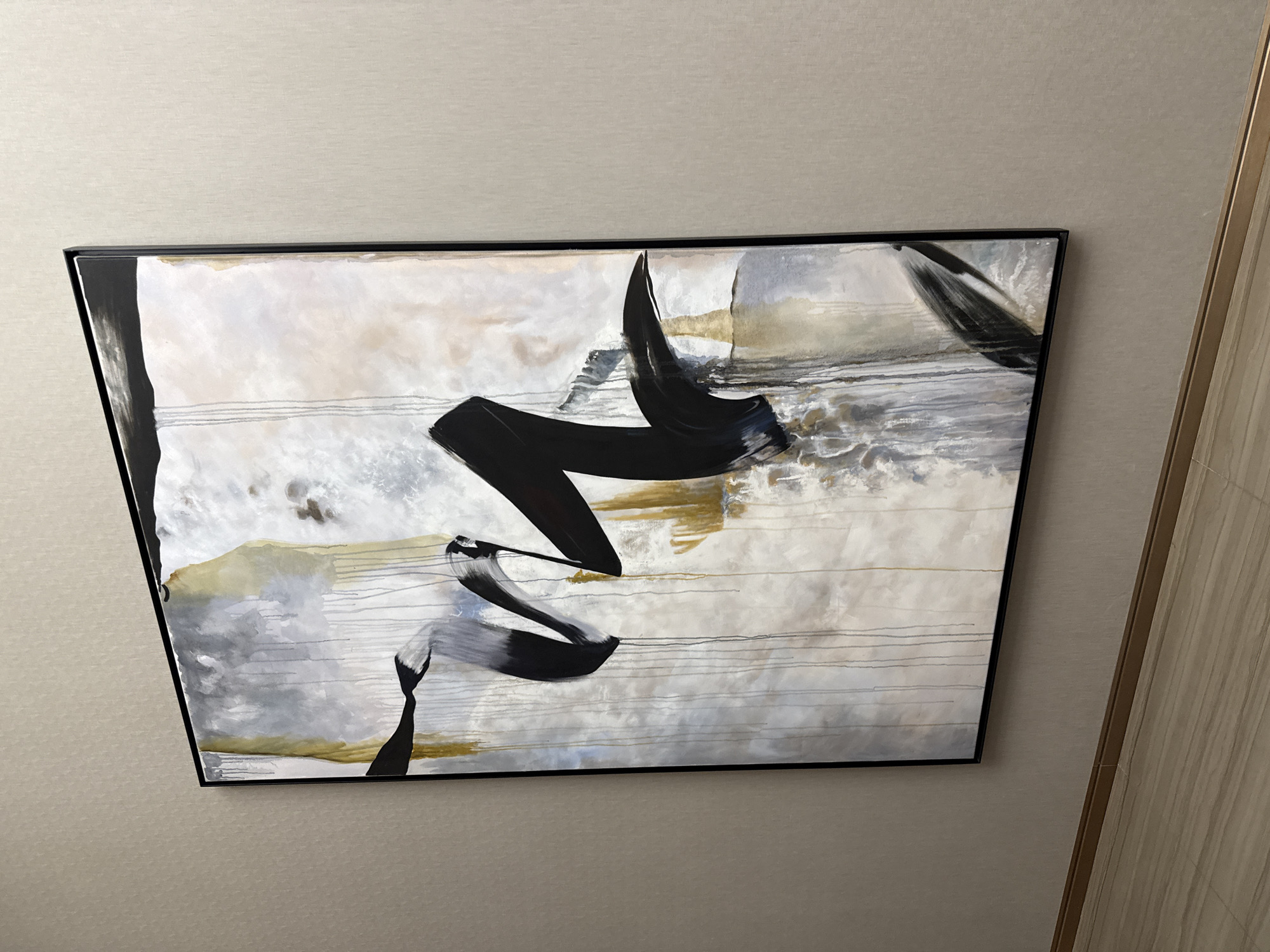 An abstract painting is centrally featured, hanging on a light beige, textured wall in an indoor setting in Khimki, Russia. The artwork is presented within a slim black frame and displays a horizontal orientation. The canvas itself is dominated by a palette of off-white, cream, and various shades of light grey, accented by subtle washes of pale yellow or gold. Bold, expressive black brushstrokes form prominent, curvilinear shapes across the composition, creating a sense of movement and contrast. Additionally, several fine, parallel horizontal lines in grey and black are visible across different areas of the painting. To the right side of the frame, a partial view of a light wood-grained panel or door is visible, running vertically. There are no people, explicit activities, or discernible text present in the image.