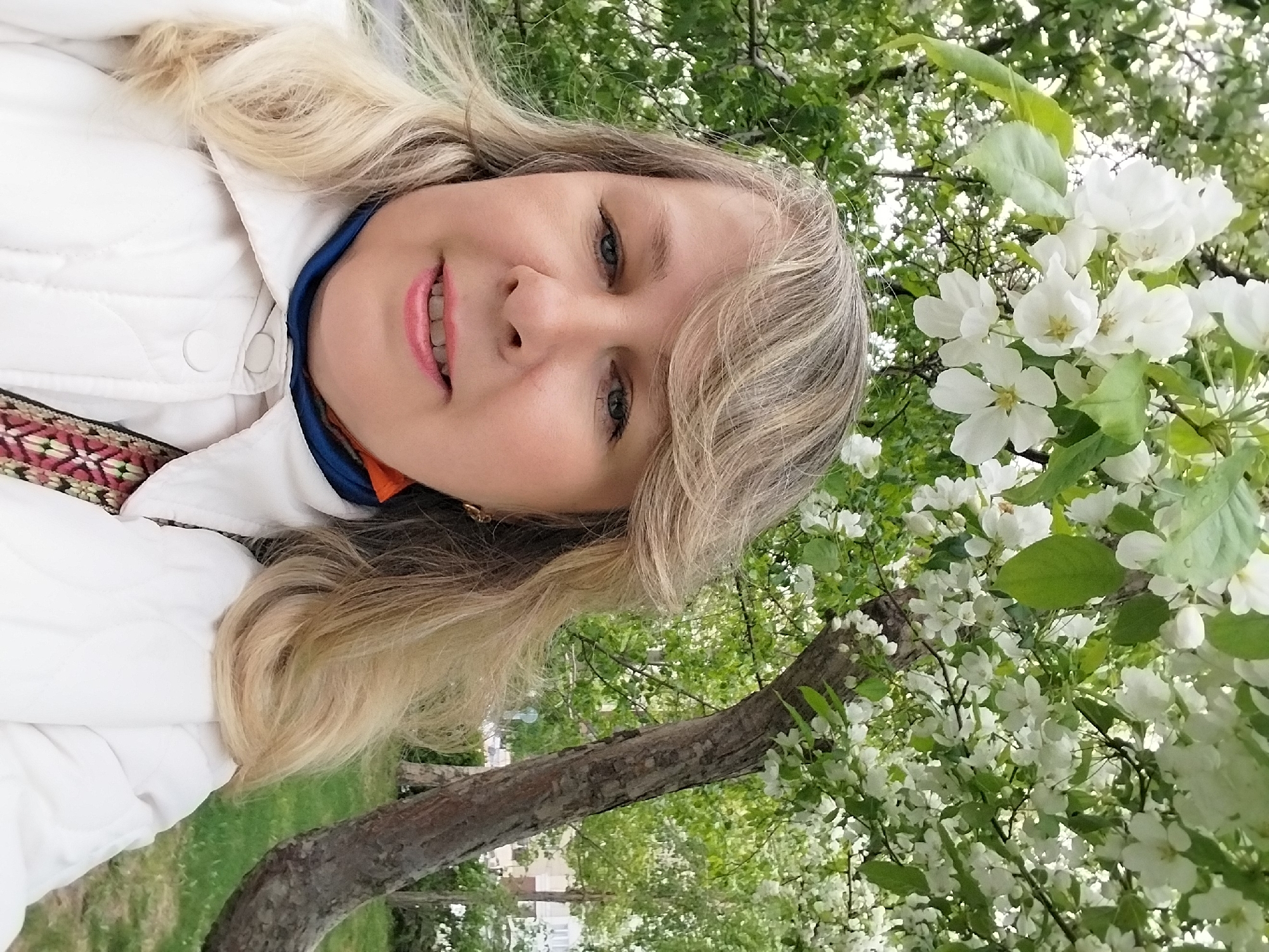 A woman with blonde hair and fair skin is posing in a selfie, with a tree branch bearing white flowers in the background. She is wearing a white jacket and a colorful patterned garment underneath. The scene appears to be outdoors in a park or garden, with greenery visible in the background. Based on information available online, the location is likely to be in the city of Orenburg, Russia.