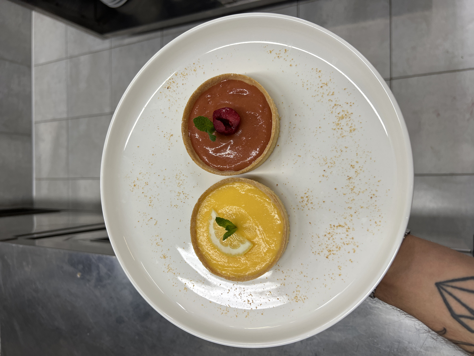 The image shows a white plate holding two small tarts. The upper tart has a light brown filling topped with a raspberry and a small sprig of mint. The lower tart has a bright yellow filling topped with a slice of lemon and a small sprig of mint. The plate is decorated with a sprinkle of what appears to be a powdered garnish. A person with a tattoo on their arm is holding the plate. The background includes a grey tiled wall and a stainless steel surface. It is likely that this image was taken in a restaurant kitchen, possibly in Budva, Montenegro.