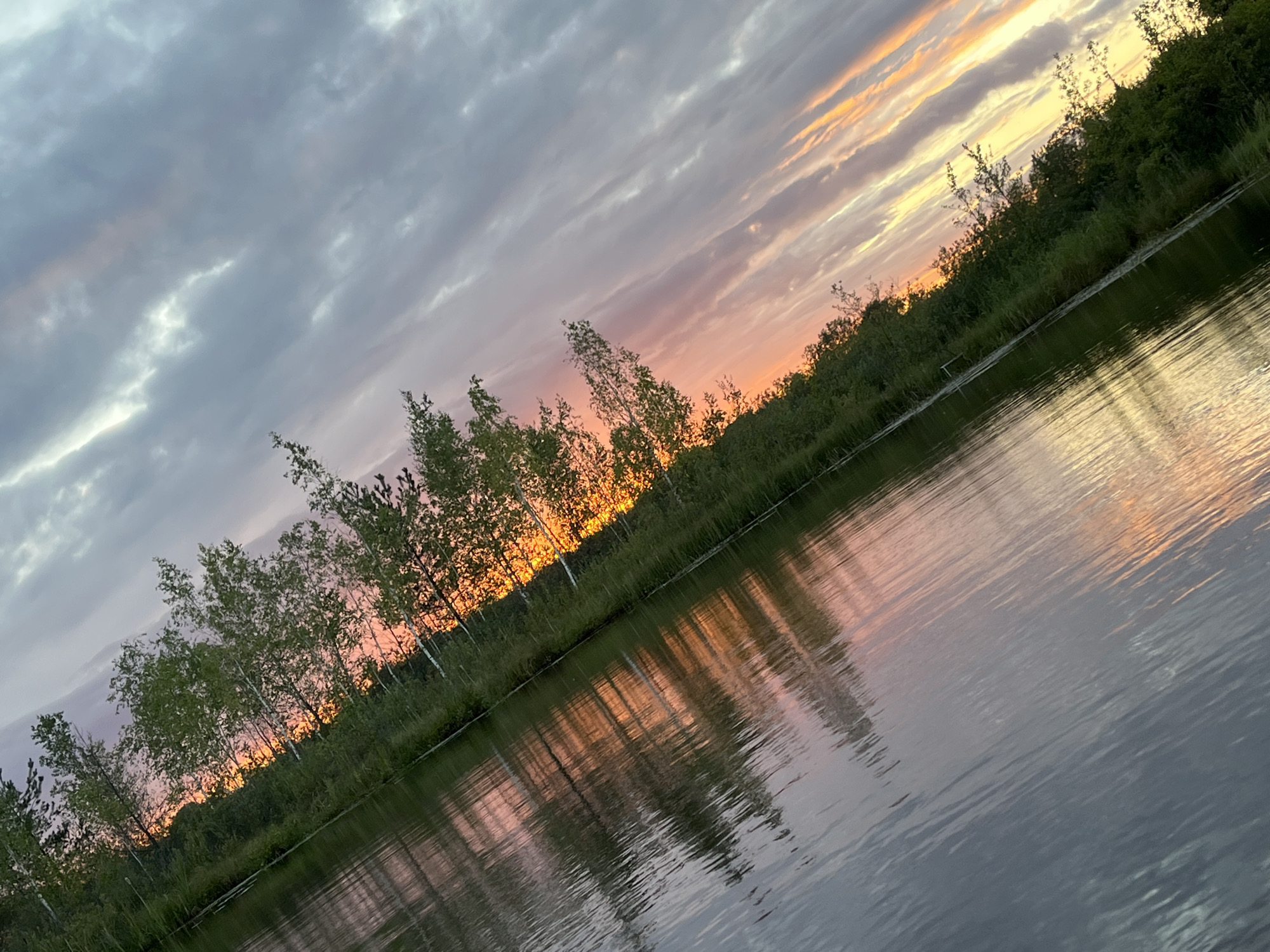The image captures a serene sunset over a body of water lined with trees. The water reflects the sky's colors, creating a mirror image of the sunset. The sky is filled with clouds, some of which are tinged with pink and orange hues from the setting sun. The trees are lush green, with their reflections extending into the water. The scene is peaceful and natural, with no signs of human activity. The photograph appears to have been taken from a low angle, possibly from a boat or the water's edge. The overall impression is one of tranquility and natural beauty, likely located in the rural landscape of Дунілавіцкі сельскі Савет, Belarus.