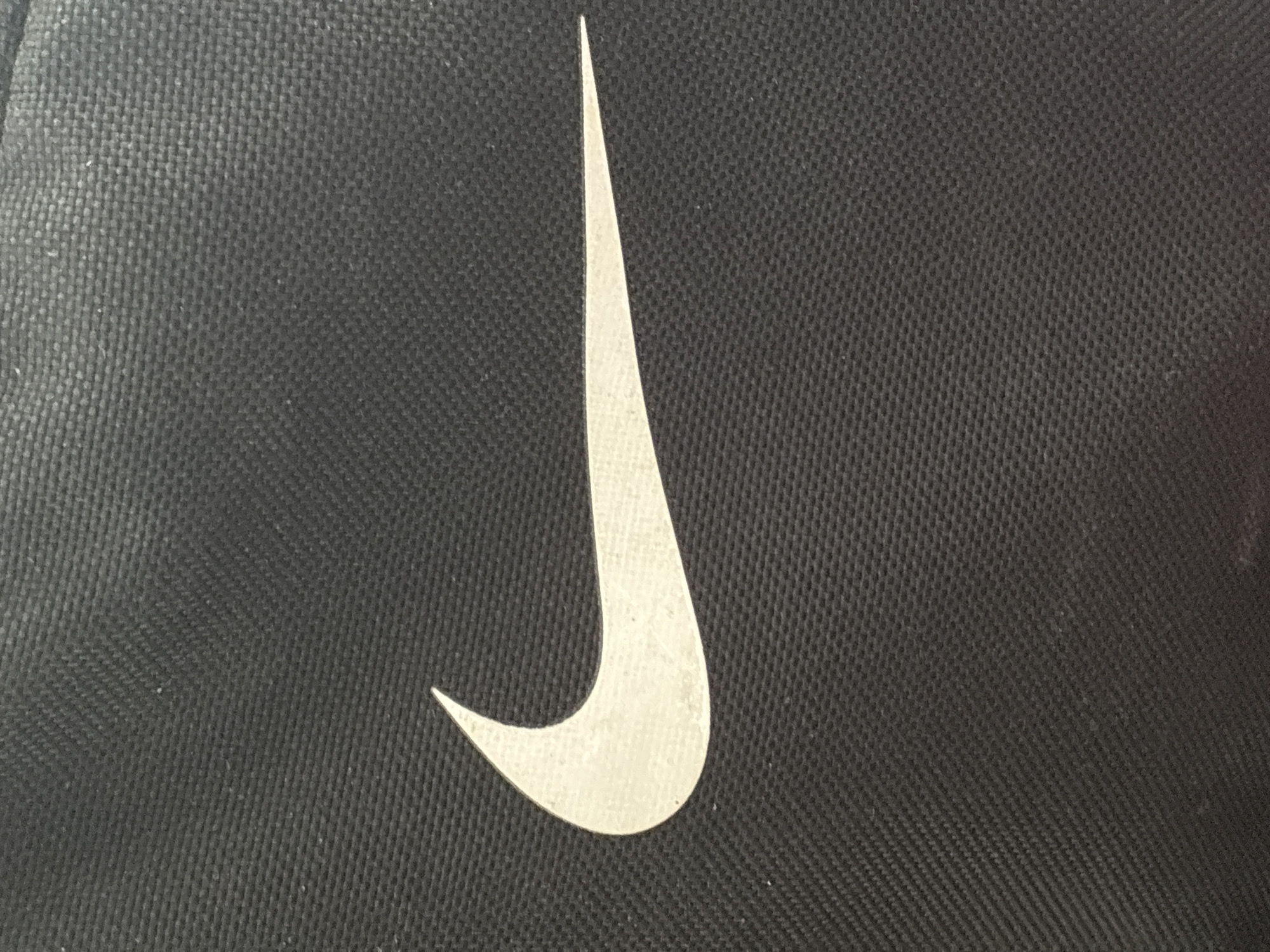 A close-up shot captures a white Nike swoosh logo against a textured black background, likely fabric. The swoosh is prominently displayed in the center of the frame, facing to the right. The black background exhibits a repeating woven pattern, characteristic of many textiles. The lighting appears to be soft, illuminating the logo without casting harsh shadows. No people, actions, or specific environmental details are visible.