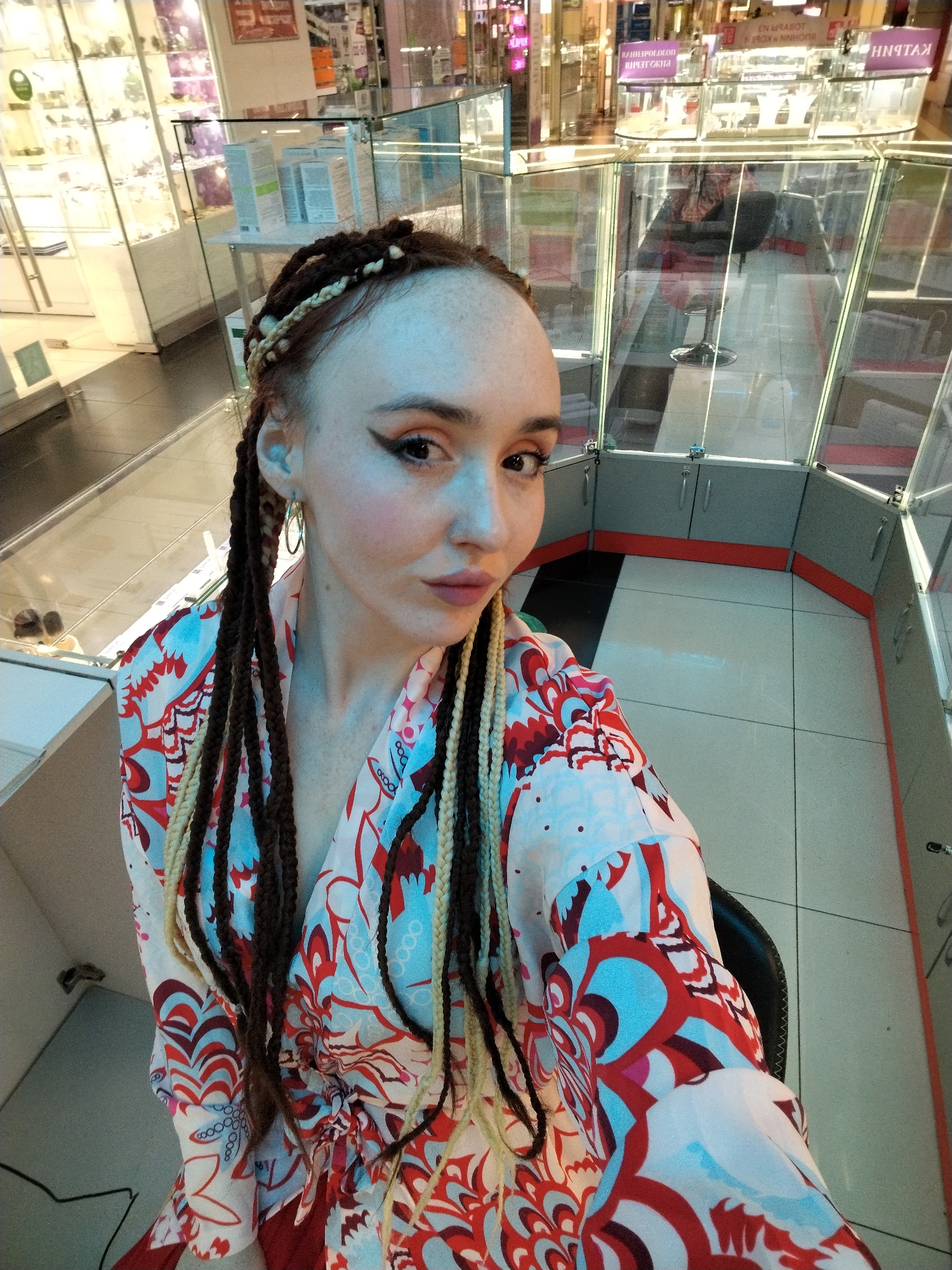 The image shows a person with light skin, wearing a colorful floral-patterned kimono-style robe. They have brown hair with lighter-colored braids, hoop earrings, and prominent eyeliner. The person is looking at the camera with a slight pout.
The background appears to be the interior of a shopping mall. Display cases containing items such as jewelry are visible. Signage in Russian is noticeable, and the architecture suggests a modern indoor shopping environment. A sign in the background says "ПЯТAЧOK" which appears to mean "Piglet".
Based on the provided information, the location is likely to be in Izhevsk, Russia.