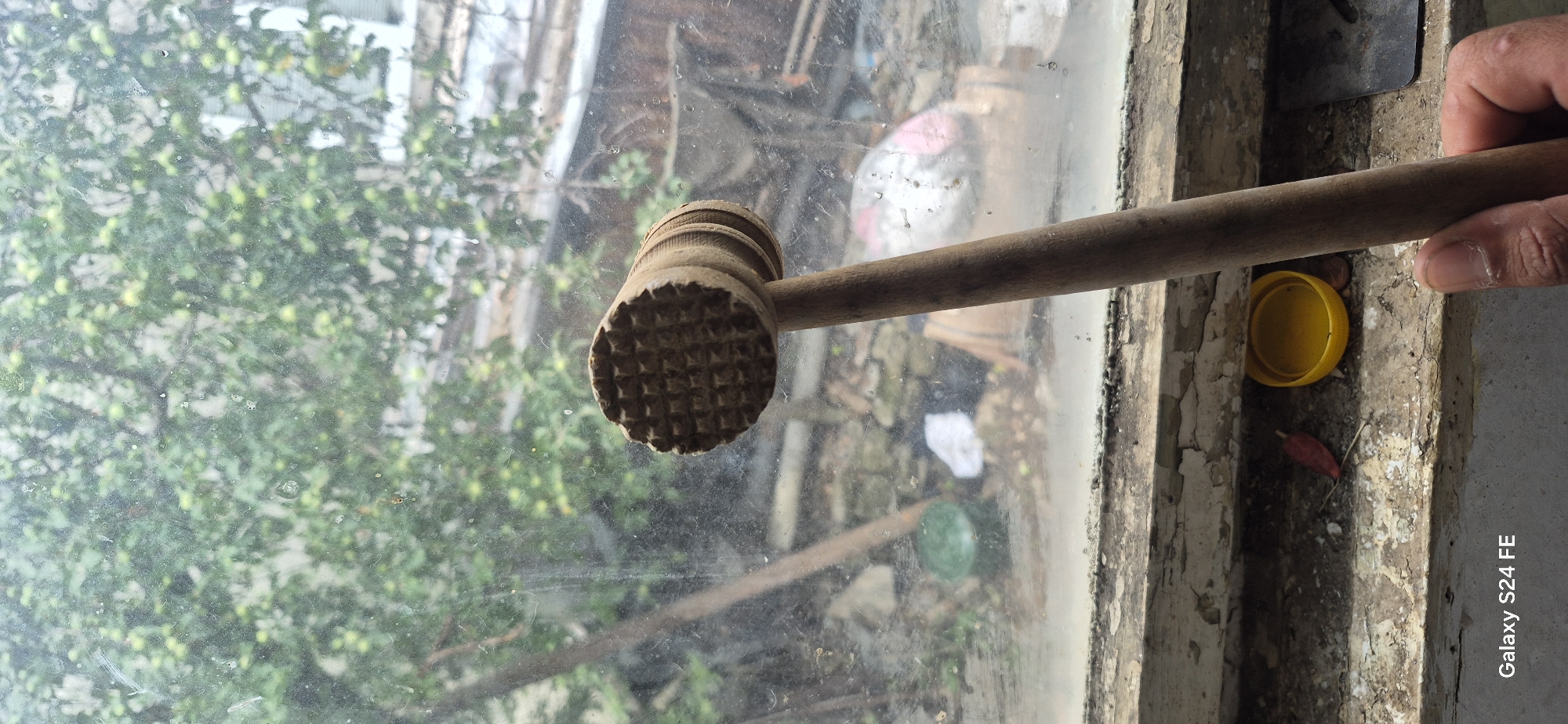 A wooden meat tenderizer with a patterned head rests on a windowsill, held by a person's hand. The tenderizer is positioned in the foreground, with its patterned head facing left. Behind it, a grimy window reveals a view of greenery and trees. The windowsill itself is weathered and peeling, with a yellow bottle cap and a red dried leaf lying on it. In the background, various objects are indistinctly visible through the window, including what appears to be a green bucket and white debris. The scene suggests an outdoor or semi-outdoor setting, possibly in a residential area. The image is tagged with "Galaxy S24 FE".