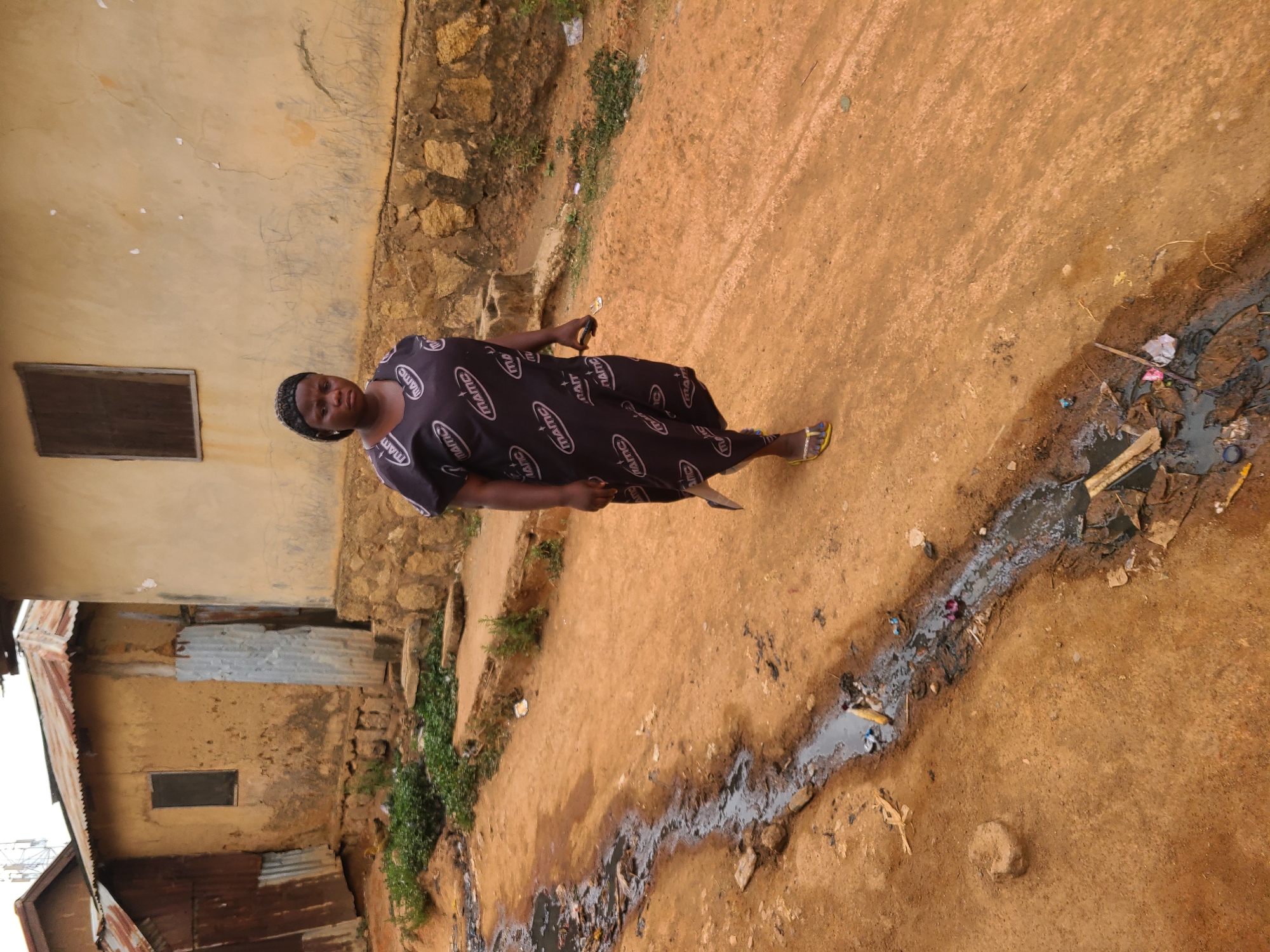 A woman stands on a dirt road in a dilapidated area of Jos, Nigeria. She wears a dark dress with white lettering and yellow sandals. A drainage ditch filled with dark water and trash runs along the edge of the road. Behind her, there are weathered buildings made of stone and corrugated metal. The sky is overcast.