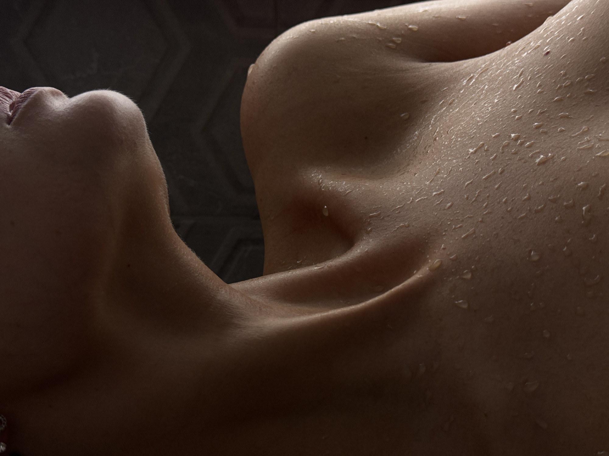 This image is a close-up, low-angle shot focusing on the shoulder, collarbone, and neck area of a person. The skin is wet, with numerous droplets of water visible. The lighting is dim, casting shadows and highlighting the contours of the body. The background is dark and appears to be a textured surface, possibly a tiled wall. There are no discernible city landmarks or elements of Chaikovsky, Russia, visible in the image. The subject's face is not fully visible, with only a hint of lips and chin in the upper part of the frame.