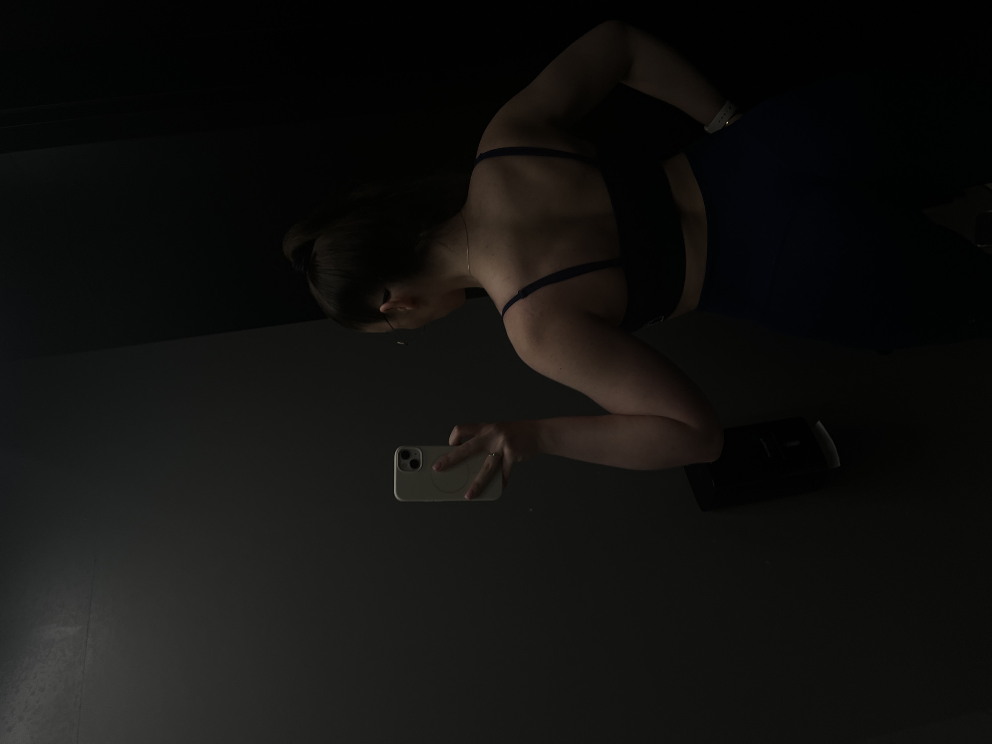 A woman takes a selfie indoors. She is wearing a dark sports bra and shorts. She holds a white cell phone in her right hand. The background is dark. A small black object is near her. Location: Unknown, Unknown.