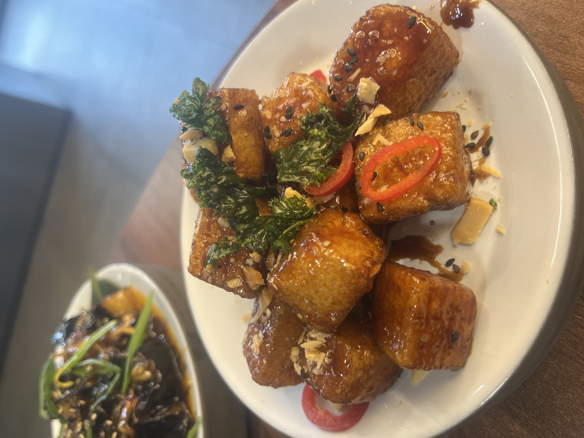 The image displays a close-up, slightly angled view of two dishes, presumably from a meal served in Sofia, Bulgaria. The primary focus is on a white, round plate piled with numerous golden-brown, bite-sized cubes generously coated in a dark, glossy, amber-hued sauce, suggesting a sweet and savory glaze. These cubes appear to be fried, possibly tofu or a starchy vegetable like sweet potato.
The main dish is elaborately garnished. Scattered among the glazed pieces are vibrant green, crispy-looking leafy herbs, possibly fried basil or kale, adding a textural contrast. Bright red chili pepper slices, cut into rings, provide a visual accent. Black sesame seeds and small, chopped pieces of what appear to be peanuts or similar nuts are sprinkled over the cubes, contributing to both flavor and crunch. Some lighter-colored flakes, possibly fried garlic, are also visible on the food. The sauce has drizzled onto the clean white plate in several spots.
In the lower-left corner, a second dish is partially visible and out of focus. It is presented in a smaller, white oval bowl and contains a dark, rich-looking liquid or sauce, garnished with thin, long green strands, likely scallions or chives.
The background is softly blurred, showing a light-colored wall or surface behind the main dish and a darker, wooden tabletop supporting the plates. The lighting appears to be bright and even, highlighting the glistening texture of the food. No people or visible text are present in the image. The scene depicts a well-prepared and appetizing culinary presentation.