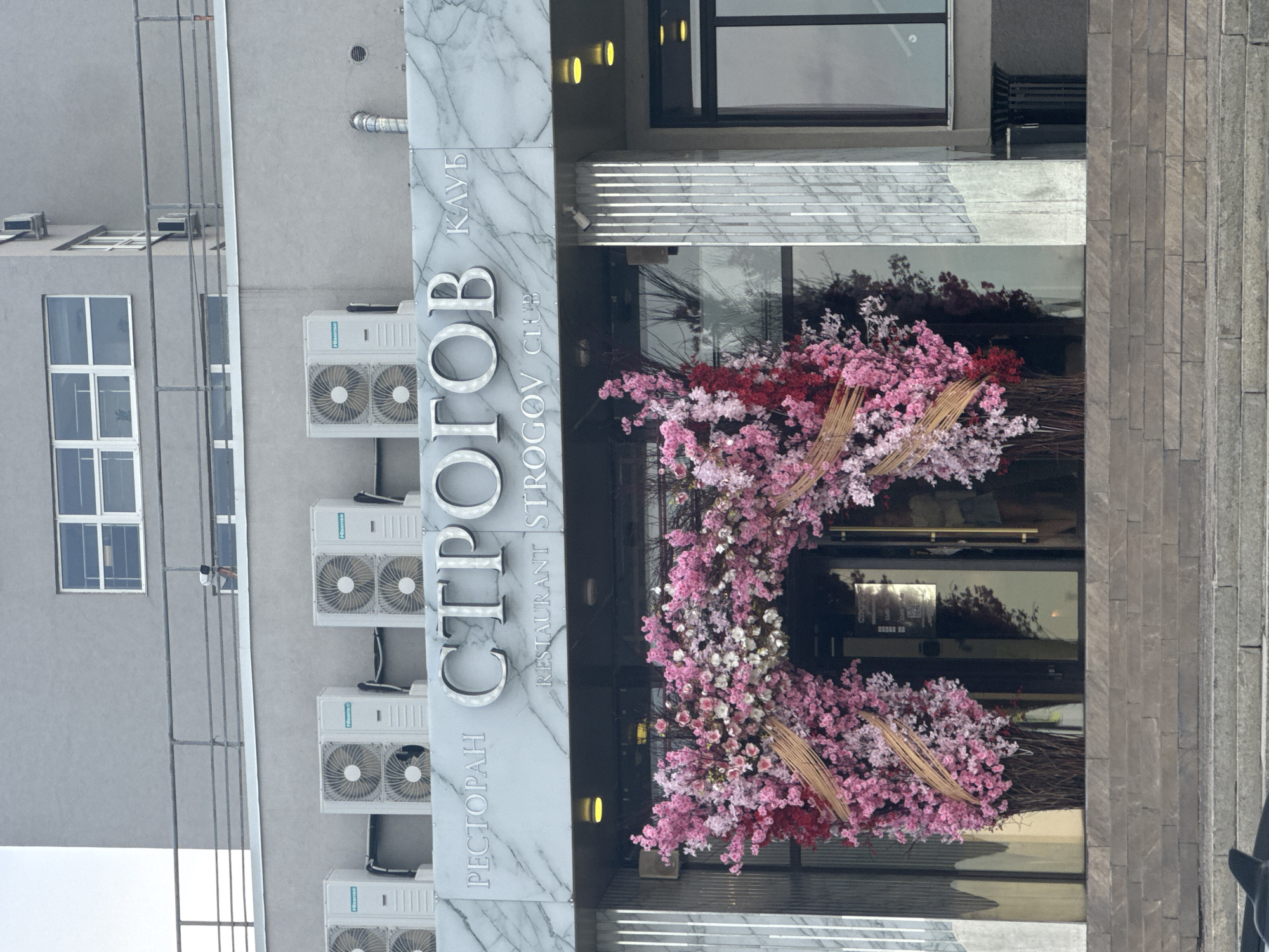 The image shows the entrance of "Stroganov Club Restaurant" in Nizhny Novgorod, Russia. The entrance is decorated with a large floral arrangement featuring pink, white, and red blossoms. The sign on the left reads "РЕСТОРАН STROGANOV КЛУБ" (Restaurant Stroganov Club) vertically. The building is gray and has multiple air conditioning units on the wall. The entrance has glass doors and some steps leading up to it. The overall aesthetic is modern with a touch of natural beauty through the floral decorations.