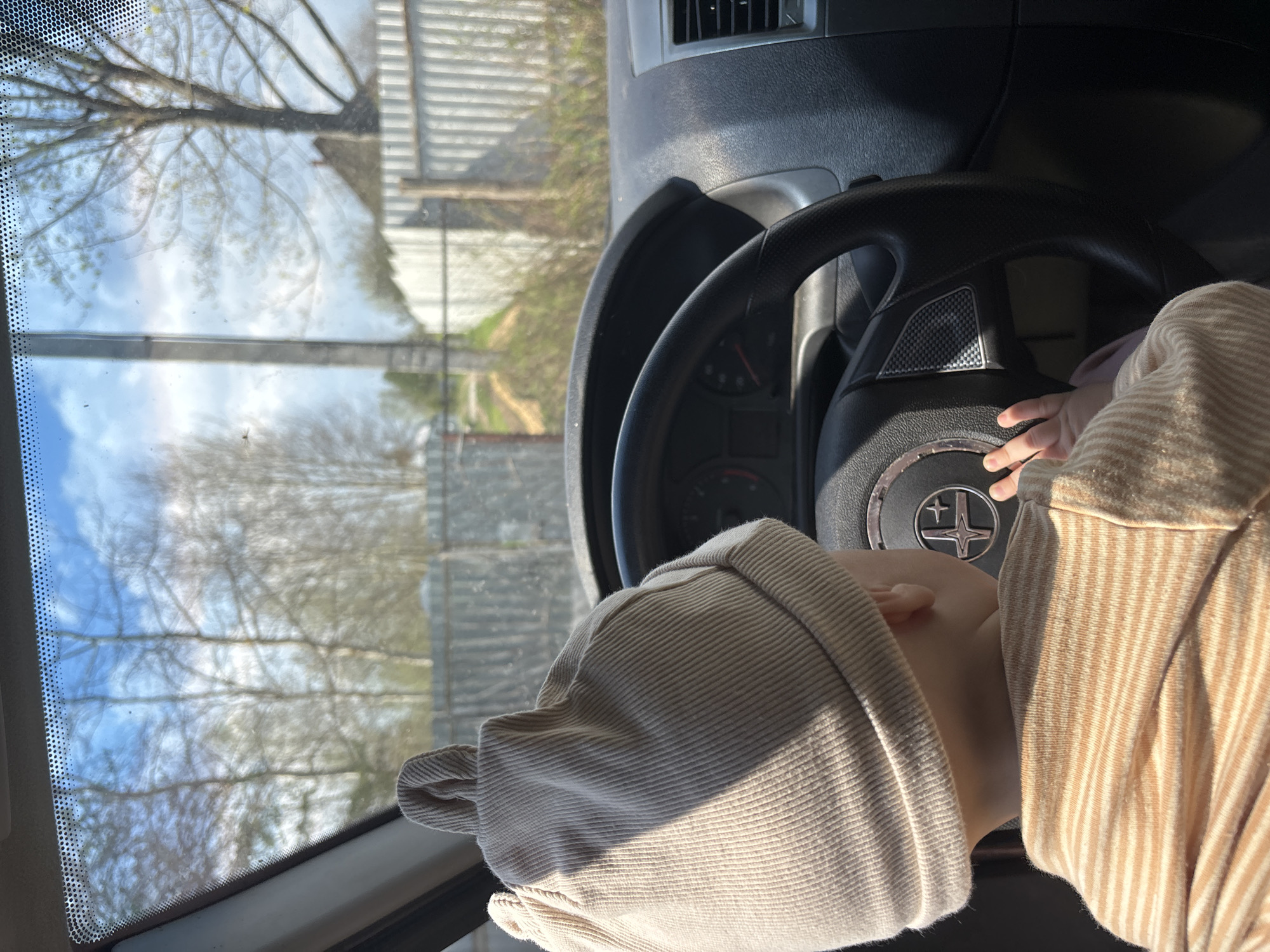 A baby wearing a beige hat and striped shirt is positioned behind the steering wheel of a vehicle. The baby's hand is near the center of the steering wheel. Outside the vehicle, trees and a partial view of a building and fence are visible through the windshield. The location is likely Мещёры, Russia based on the request.