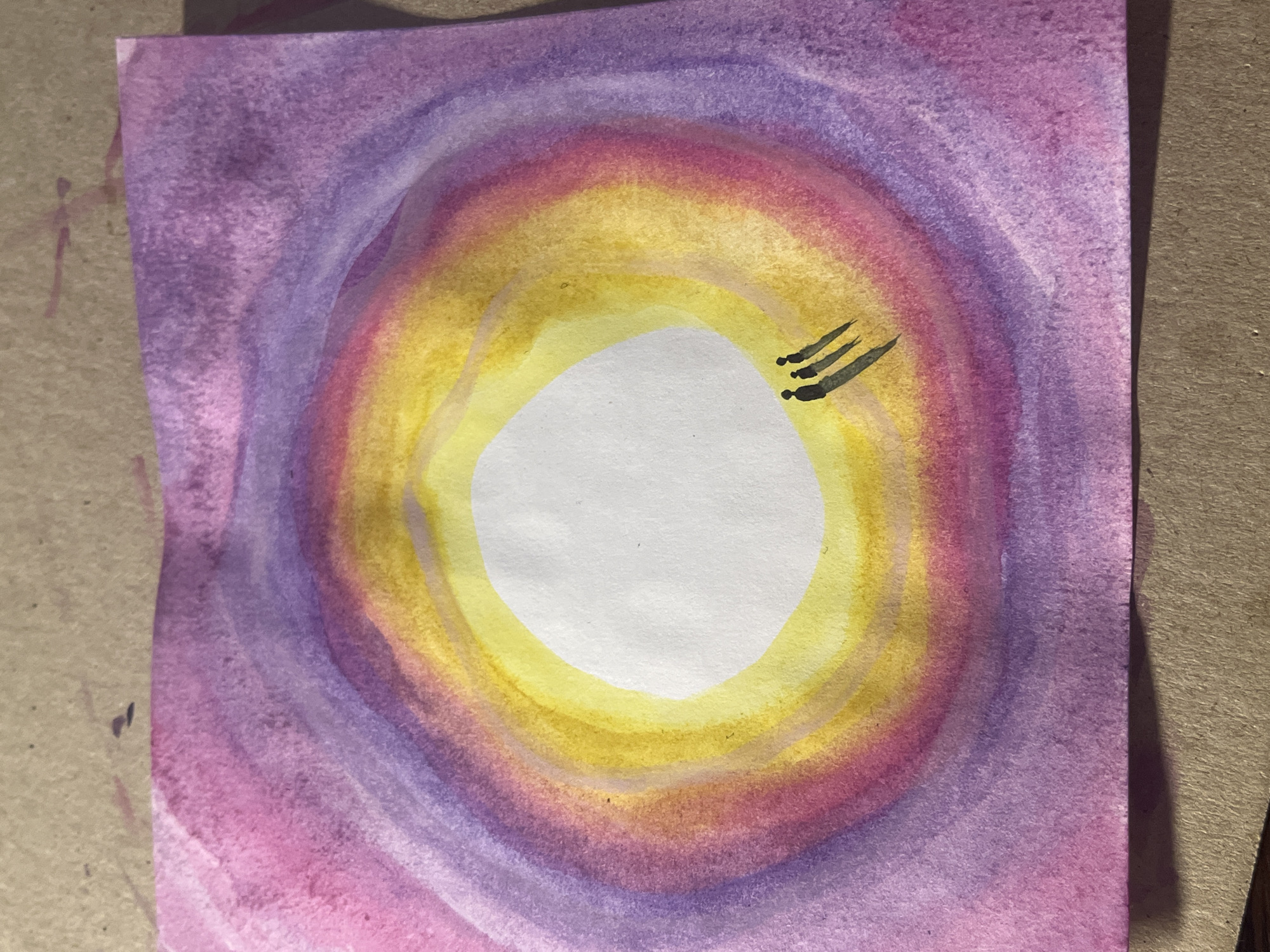 The image shows a watercolor painting on a piece of paper. The central motif is a concentric pattern of colors surrounding a white, roughly oval-shaped space in the center. The colors transition from purple/pink on the outer edges to yellow nearer to the center. Three short, dark lines, angled towards the center, are present in the yellow area. The background on which the painting rests is a neutral brown color. The scene is not the city of Saint Petersburg, Russia.