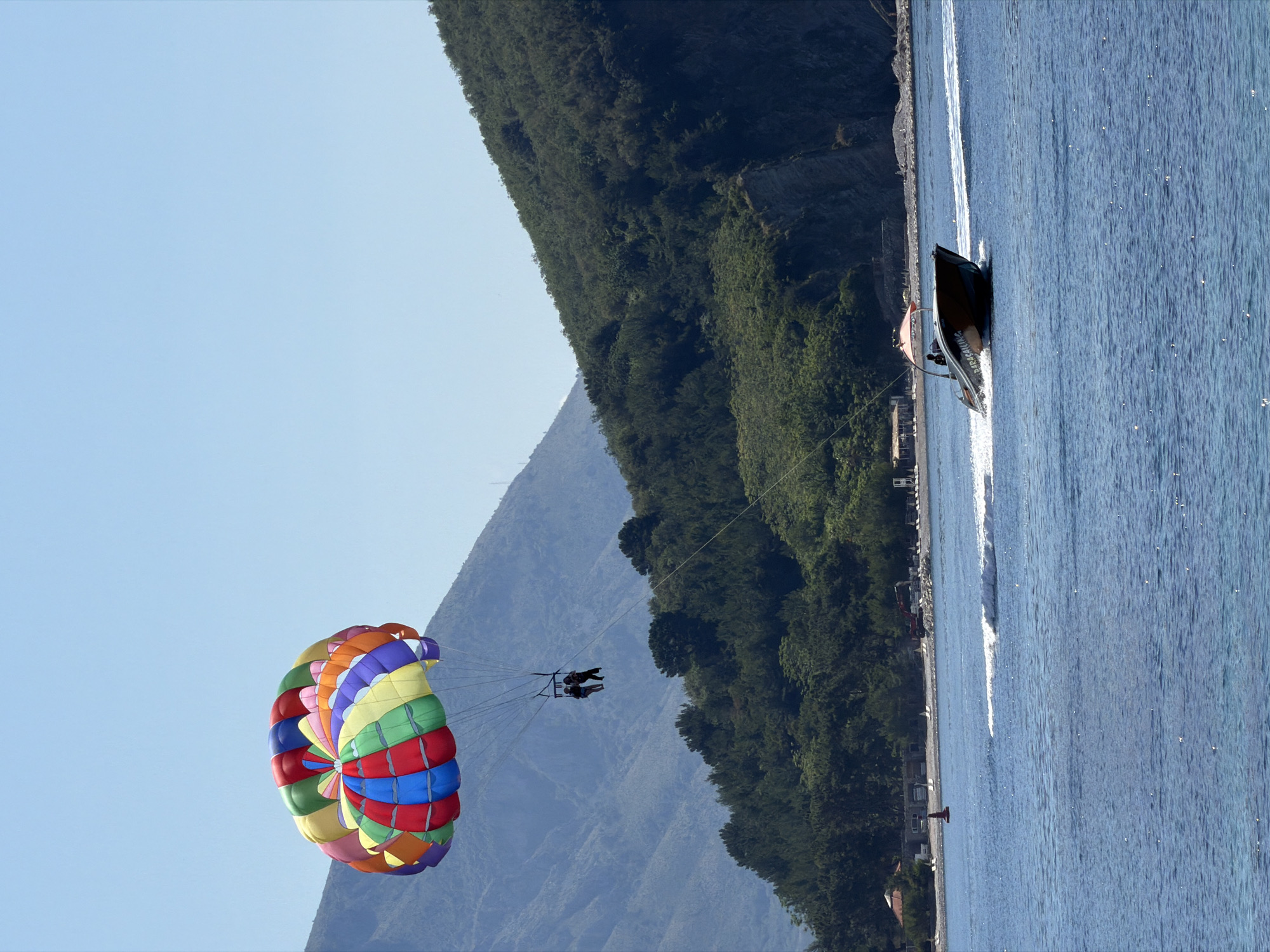 This image captures a bright, sunlit coastal scene identified as Budva, Montenegro, depicting parasailing activity. The upper portion of the frame is dominated by a clear, light blue sky.
In the upper left quadrant, a large, fully inflated parasail is aloft, displaying a vibrant multi-colored canopy segmented in hues of red, blue, green, yellow, orange, purple, and pink. Suspended directly below the parasail are two individuals in a harness, appearing as dark silhouettes against the distant landmass. A long tow line extends diagonally downwards from the parasailers towards the lower right of the frame, connecting them to a moving boat.
The lower right quadrant features a dark-hulled motorboat, likely a purpose-built parasail boat, with the word "PARASAILING" visible on its side. The boat has an orange canopy over its stern and is actively moving from left to right, generating a distinct white wake behind it. An operator is visible at the helm.
The background consists of a layered landscape of mountains. To the left, distant, hazy blue-grey mountains rise under the clear sky. Closer to the right and center, a steep, lush green mountain, densely covered in trees, slopes down to the sea. Rocky cliffs are visible at the base of this green mountain.
Along the coastline at the base of the green mountain, a narrow stretch of beach is discernible, featuring scattered white lounge chairs and several low-rise buildings or structures. A red excavator or similar construction vehicle is present on the beach in the mid-distance. A red navigational buoy floats in the water near the distant shore. The sea in the foreground and mid-ground is calm, displaying a deep blue hue with subtle reflections of sunlight.