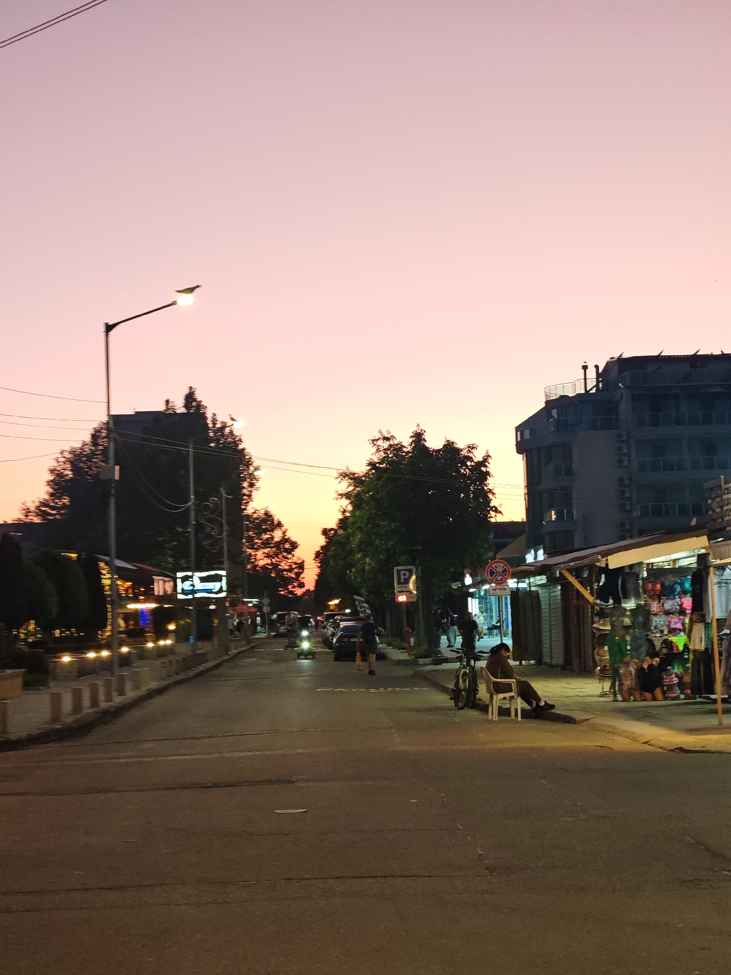 Captured at dusk in the city of Nesebar, Bulgaria, the image presents a street scene. The sky transitions from pink to orange as the sun sets. The street is lined with buildings, some of which are lit, and various trees. On the right side of the road, vendors have set up stalls displaying goods, and a person is seated on a chair by the curb. A bicycle is parked next to them. Further up the street, pedestrians are walking and cars are moving. A street lamp stands tall on the left, illuminating the area. The scene is quiet, possibly depicting a typical evening in the town.