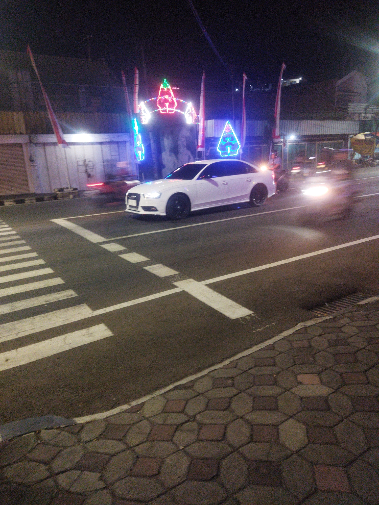 A white sedan is stopped at a pedestrian crossing on a city street at night. The car is a modern model with sleek lines and black wheels. The street is paved and has white lane markings. Behind the car, there are some colorful lights that appear to be decorations. The surrounding environment includes buildings and flags. The image is taken from a low angle, looking up at the car and the street.