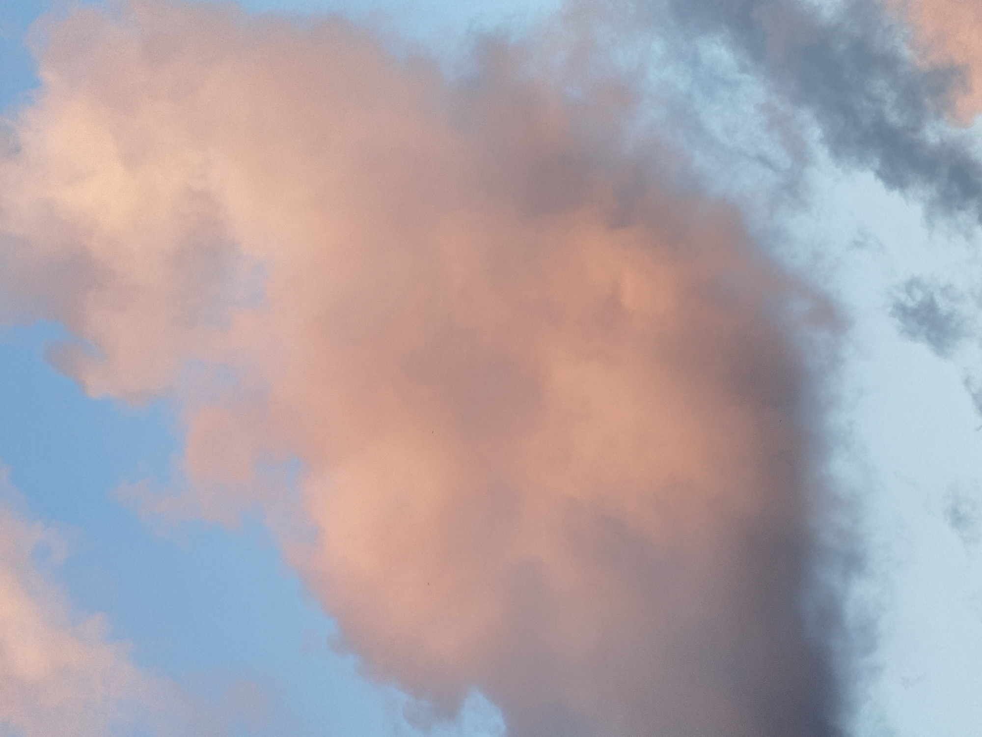 The image is a close-up shot of a sky filled with clouds, bathed in the warm hues of sunset. The sky transitions from a pale blue to soft oranges and pinks where the sunlight is directly hitting the clouds. In contrast, some clouds on the right and bottom of the frame appear darker, with shades of gray and blue, suggesting they are further from the direct light source or are denser. There are no discernible human subjects, objects, or actions within the frame, and thus no interactions. The environment is entirely the sky. The image does not provide any contextual information about the city of Kuta, Indonesia, or any specific location within it.