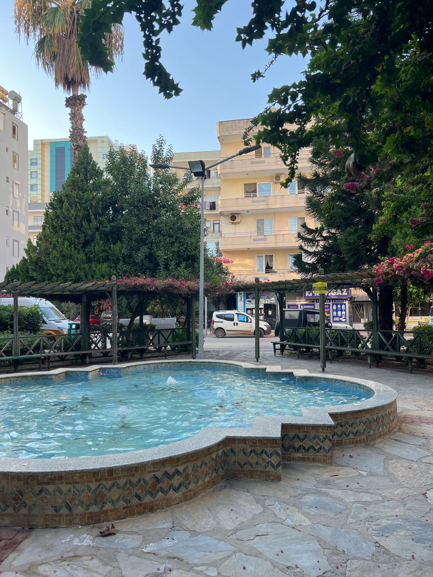 The image depicts a public fountain area in Alanya, Turkey, with an urban background. In the foreground, a kidney-shaped fountain filled with clear blue water is the central focus. The fountain's decorative base features a mosaic pattern of diamond shapes in brown, tan, and dark blue, topped with a granite-like coping. The surrounding ground is paved with irregular, light-colored flagstones, with some scattered pink flower petals.
Behind the fountain, a wooden pergola with benches is visible, providing seating. This area is bordered by a low wooden fence. Beyond the pergola, there are several trees, including a tall palm tree on the left and dense evergreens and flowering trees with pink blossoms on the right.
The background consists of multi-story buildings. The closest building to the right is a light yellow, several-story structure with balconies and windows, and air conditioning units visible. To the left of this, another building is partially visible, with a facade featuring light blue and yellow sections and numerous windows. A street or sidewalk runs in front of the buildings, with parked vehicles including a white van and a white car. Signs are visible on the shops behind the pergola, with text including "TEKNIK," "IGAZ," and "KONGAZ." The sky above is clear and blue, indicating daytime. The overall scene suggests a pleasant outdoor public space in a city.