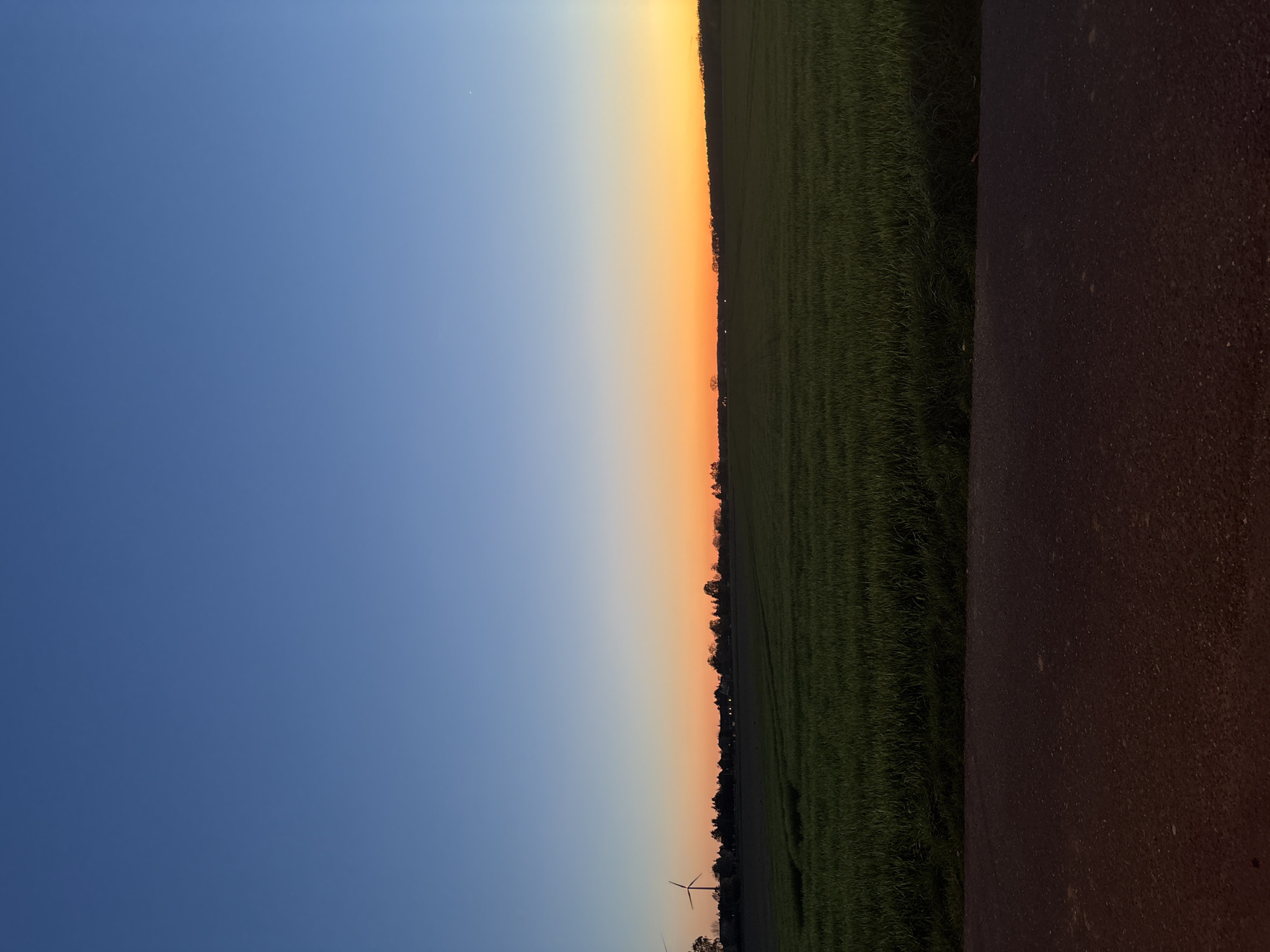 The image depicts a landscape in Wegberg, Germany, at dusk. The sky transitions from a deep blue on the left to orange and yellow hues on the right. A field of green crops stretches across the middle of the image, bordered on one side by trees. A paved road occupies the rightmost part of the frame. In the distance, wind turbines are visible on the horizon.