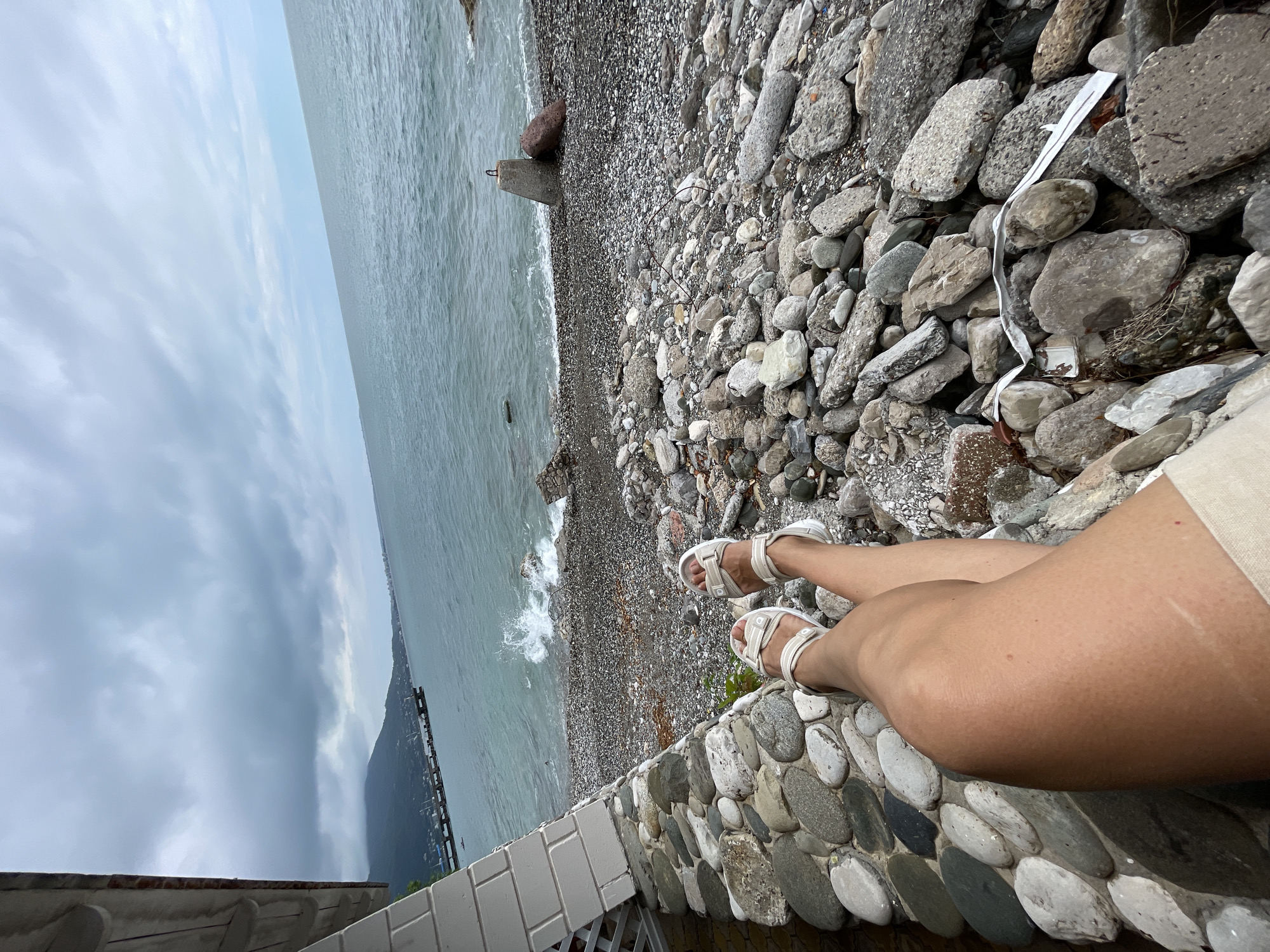 The image shows a person's legs crossed at the knees, wearing white sandals. The person is sitting on a wall made of rounded stones. In the background, there is a pebble beach with a stormy sea and a cloudy sky. A pier with boats can be seen in the distance. The location is the city of Abaata, Abkhazia. The overall scene suggests a seaside relaxation on a gloomy day.