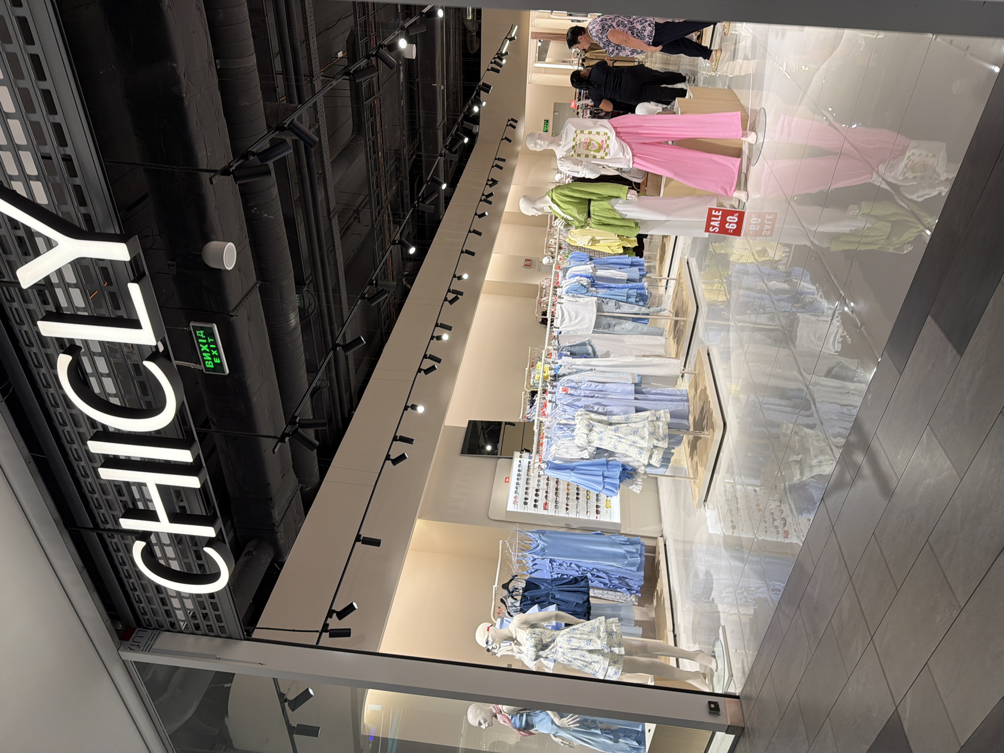The image shows the exterior of a clothing store called "CHICLY" located in Kyiv, Ukraine. The store has a large, illuminated sign with the store's name. Inside, mannequins are displayed wearing various outfits, and racks of clothing are visible. The store appears to be in a shopping mall, with a tiled floor that reflects the lights and merchandise. Two women are seen browsing in the store. The overall scene depicts a retail environment with a focus on fashion.