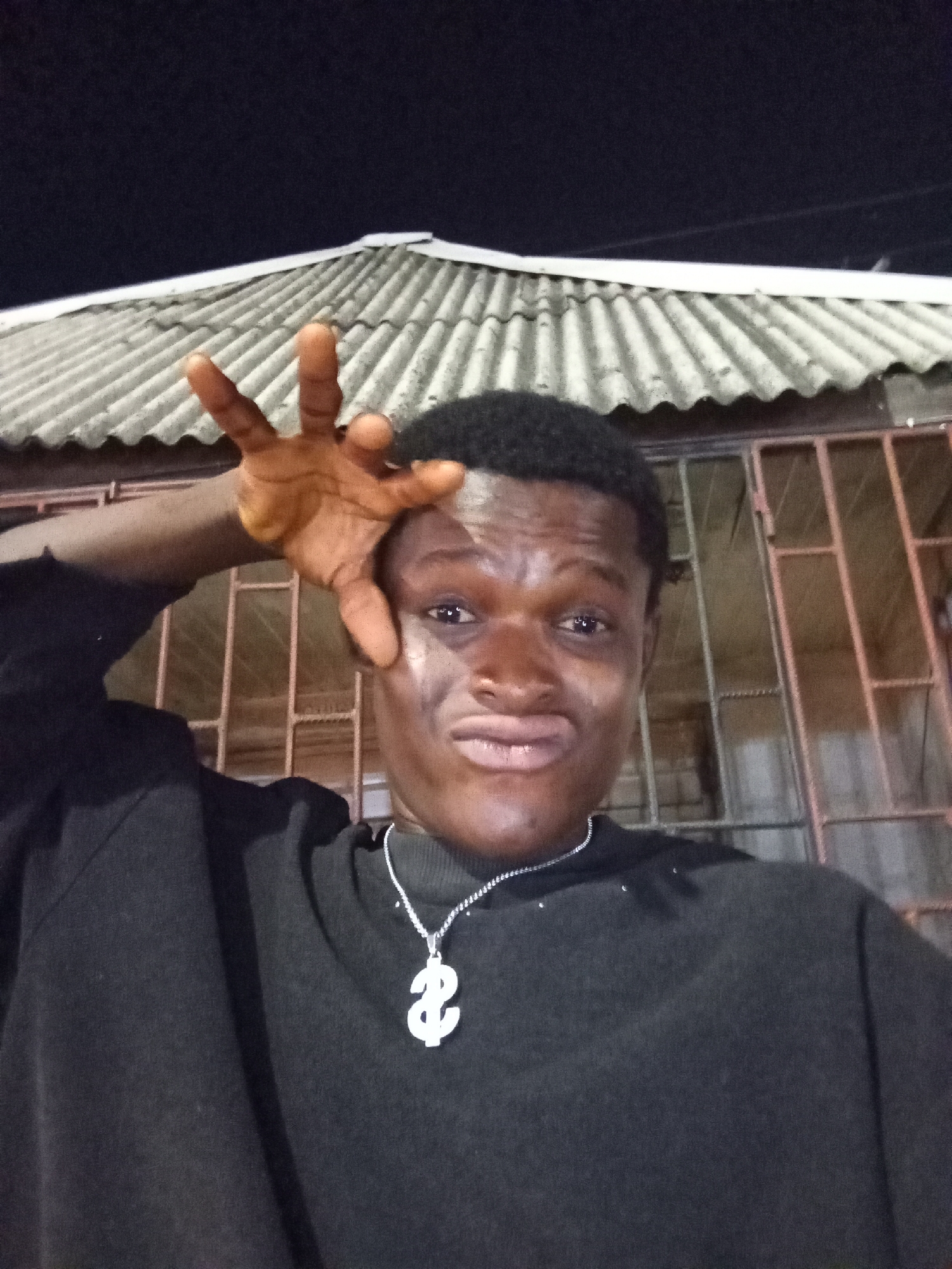 A medium shot shows a young Black man with dark hair standing outside at night. He is wearing a dark-colored, long-sleeved shirt and a silver chain with a dollar sign pendant. He is holding his left hand up near his head, fingers spread, and making a silly face with puffed-out lips. Behind him is a building with a gray corrugated metal roof and a barred window or gate. The location is in Ado, Nigeria.