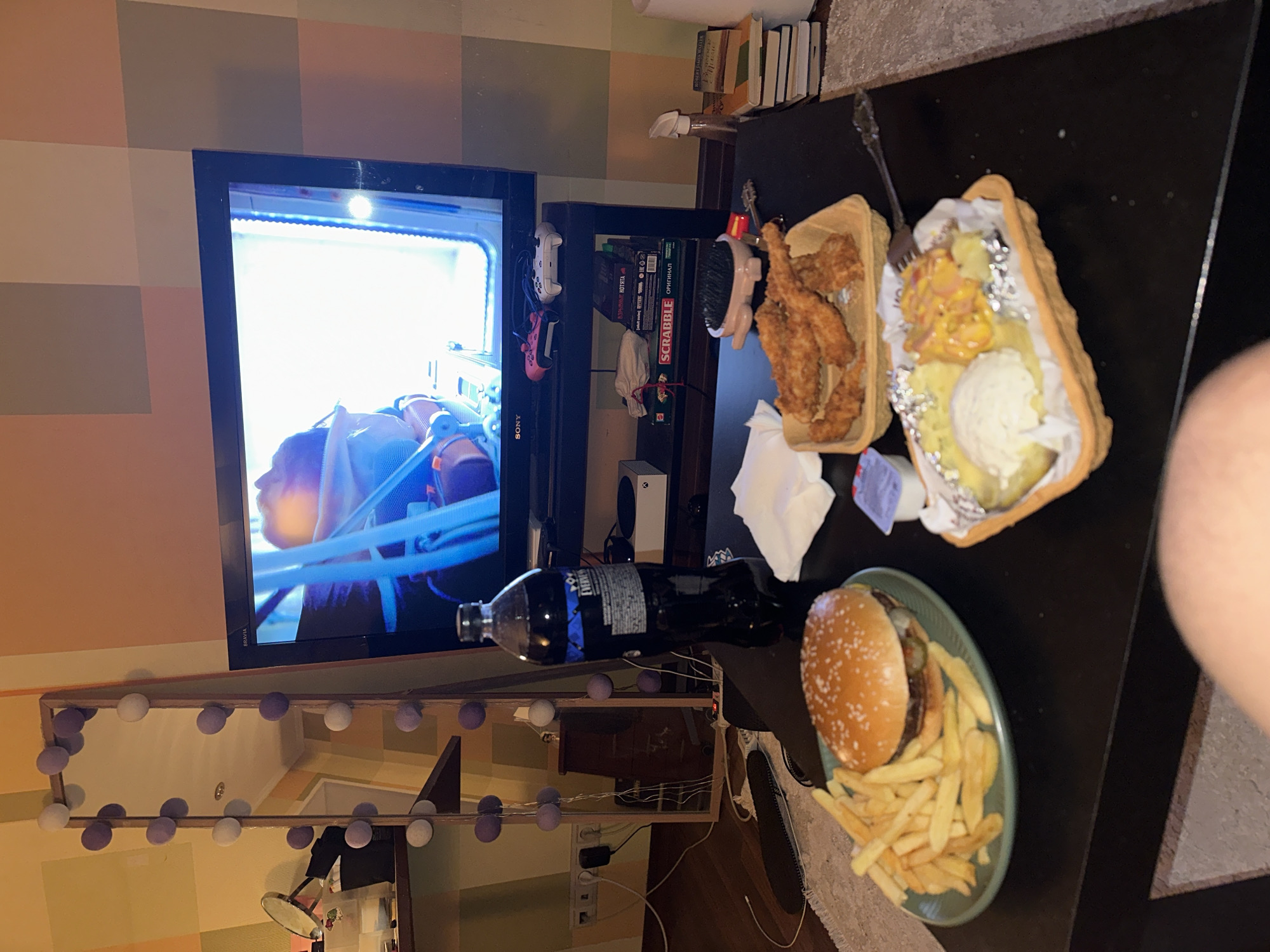 A person is watching television while having fast food. The TV is on a stand with game consoles and a Scrabble game. On a black table in front of the TV, there is a plate with a burger and fries, a container with fried chicken, and a container with a baked potato. A bottle of dark liquid is on the table. A mirror with lights is below the TV. The walls have a pattern of horizontal stripes. The location is not Moscow, Russia, and the image contains no identifiable information that would suggest the location.