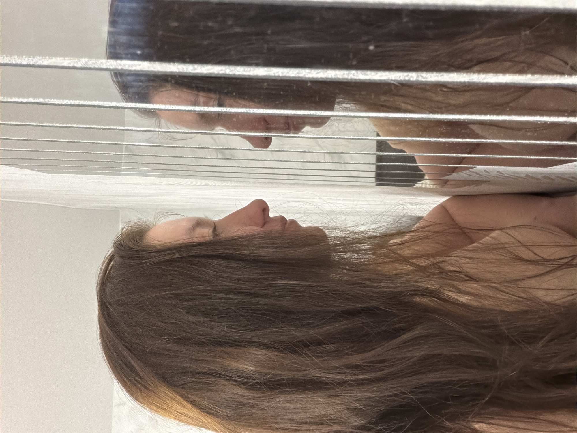 A woman lies on her side with long brown hair flowing across her shoulder and chest. Above her, her reflection is seen in a mirror behind Venetian blinds. The background is plain and white. The location of Tokkari, Russia, is not apparent from the image.