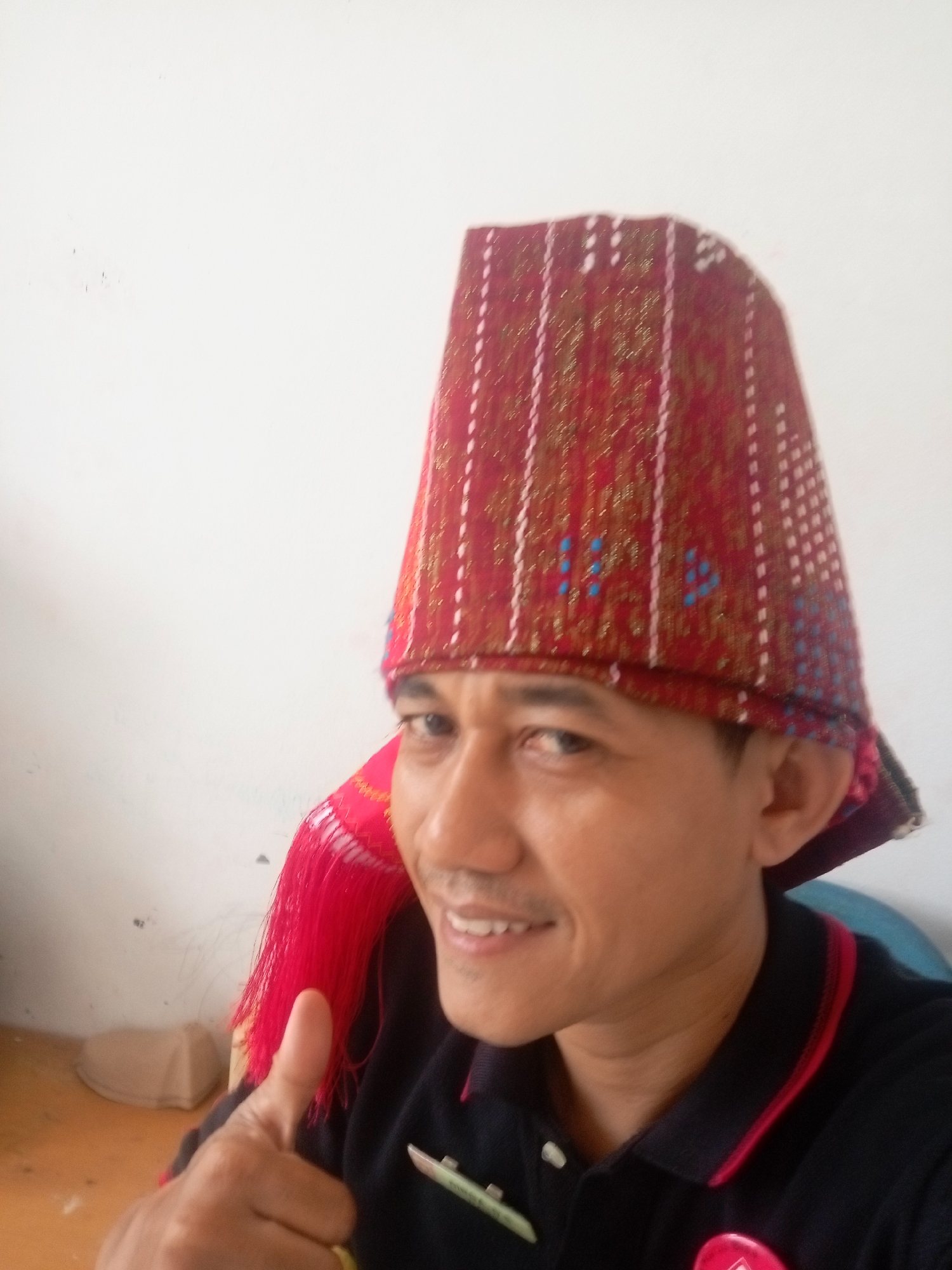 A man is posing for a selfie, wearing a red and gold patterned conical hat with red tassels. He is smiling and giving a thumbs-up. He is wearing a dark blue polo shirt with red trim. The background is a plain white wall and a light brown wooden surface. The image is likely taken indoors, possibly in Batam, Indonesia, given the traditional headwear.