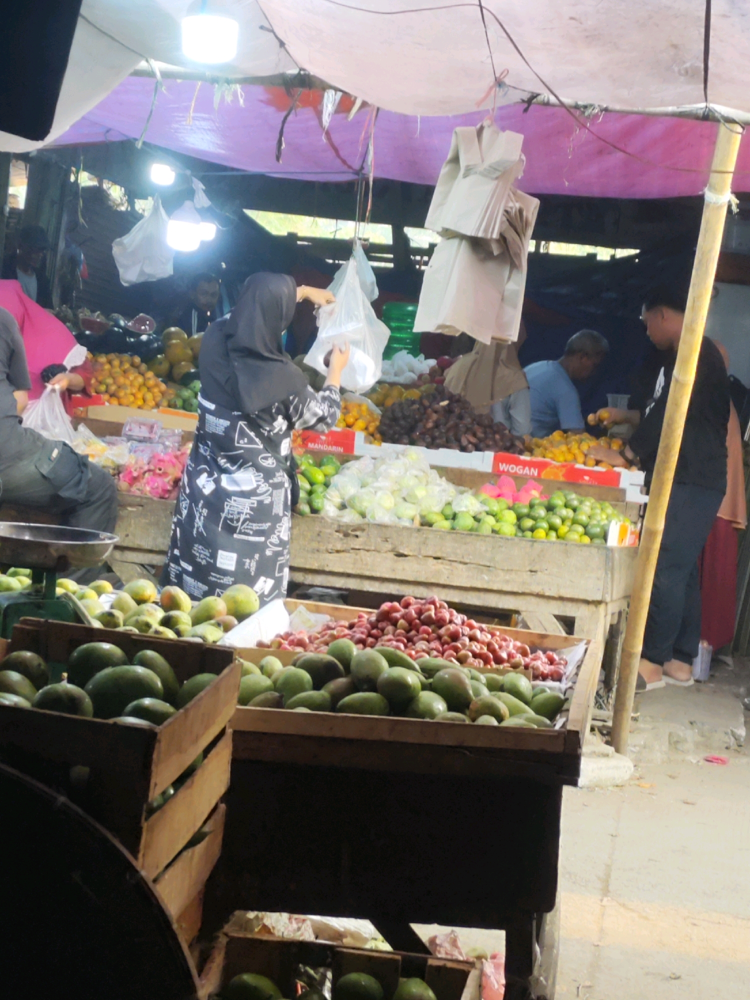 The image depicts a bustling outdoor market scene in Karawang, Indonesia, likely a fruit and vegetable stall. A woman wearing a black hijab and a patterned apron is seen actively bagging produce, suggesting she is a vendor. In the background, other individuals are visible, including a man in a blue shirt and another in a black shirt, likely customers or fellow vendors. Various fruits and vegetables are prominently displayed in wooden crates and on tables, including oranges, limes, and what appear to be avocados and tomatoes. The market is covered by a temporary tarp, supported by bamboo poles, and illuminated by hanging lights, indicating it is likely an evening or covered daytime market. The ground appears to be a dirt or paved surface, common in such marketplaces.