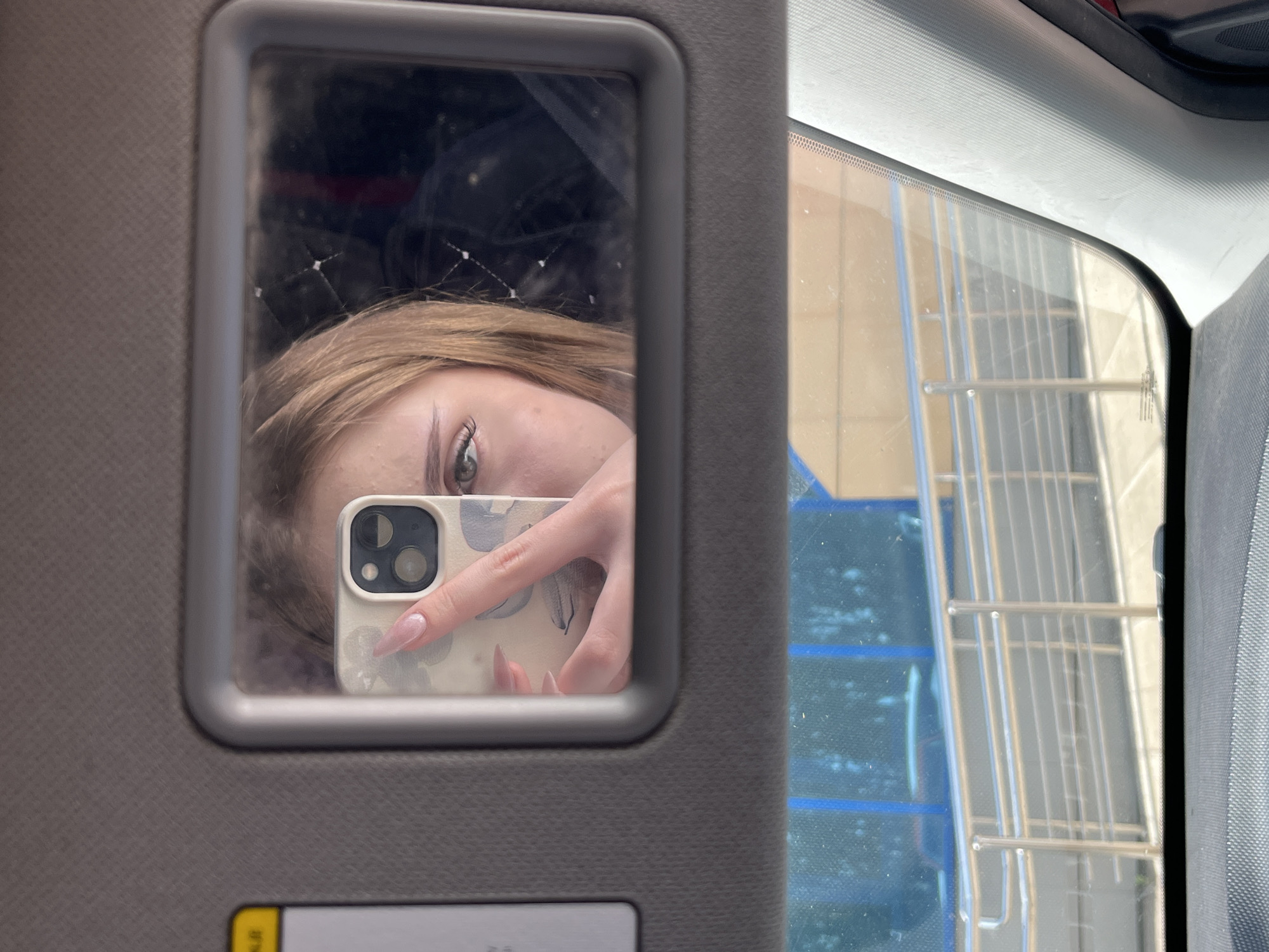 The image is a selfie taken in a car's visor mirror. The reflection shows a young woman holding a smartphone. Her face is partially visible, with one eye and a section of her forehead and cheek in focus. She has blonde hair and is wearing makeup, including false eyelashes. Her right hand is visible, holding the phone, and she has long, light pink fingernails. The background of the mirror reflection is dark and indistinct, with some faint linear patterns suggesting a textured surface or reflections. Below the visor mirror, the car's windshield shows a reflection of the exterior environment, featuring a building with blue accents, metal railings, and a walkway. The car interior is visible at the bottom of the image, including the dashboard and a portion of the A-pillar. The image appears to be taken during the daytime, with sunlight casting shadows and highlighting textures.