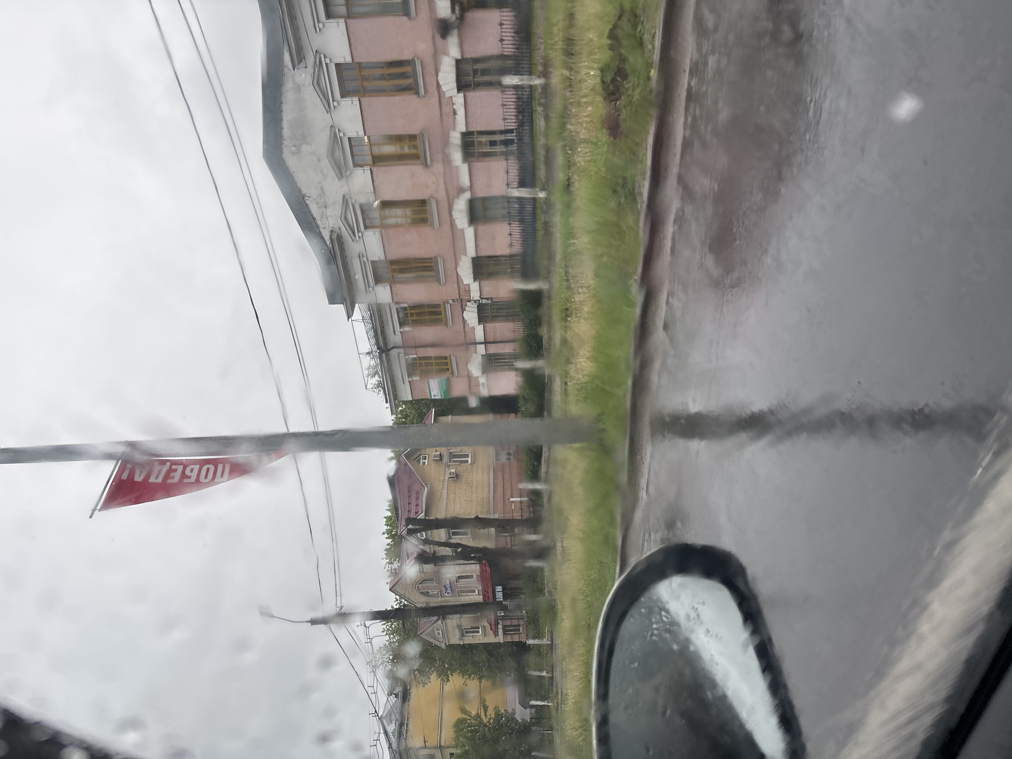 The image depicts a view from inside a vehicle, likely a car, on a rainy day. A blurred side mirror is visible in the lower right corner. The windshield is covered in raindrops.
Outside, there is a street in what appears to be a residential area. On the left side of the street, there is a multi-story, pink building with multiple windows. Adjacent to it are smaller buildings and trees.
A red banner with white Cyrillic text (likely "ПОБЕДА" meaning "Victory") is attached to a pole crossing the street. Electrical wires are also strung across the street.
The road surface appears wet, reflecting the buildings and sky.
Based on the context provided (Rusanovo, Russia), the architecture and Cyrillic text are consistent with a Russian urban environment.
