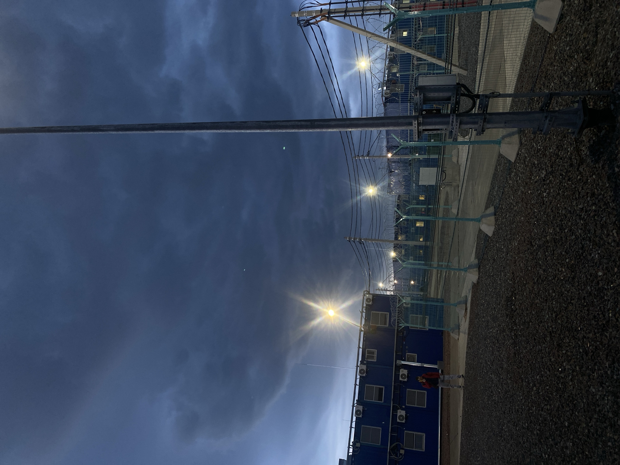 The image shows an exterior view at dusk, possibly in an industrial or semi-urban setting in Koskolovo, Russia. A long, gray pole extends from the lower right to the upper left of the frame. A series of blue, modular buildings with visible air conditioning units are on the right side of the image. A person wearing a red jacket and light-colored pants is standing near the base of the buildings. Streetlights illuminate the scene, and dark, cloudy sky is visible above. A chain-link fence runs along the right side, adjacent to a gravel surface. Power lines are strung across the top portion of the image.