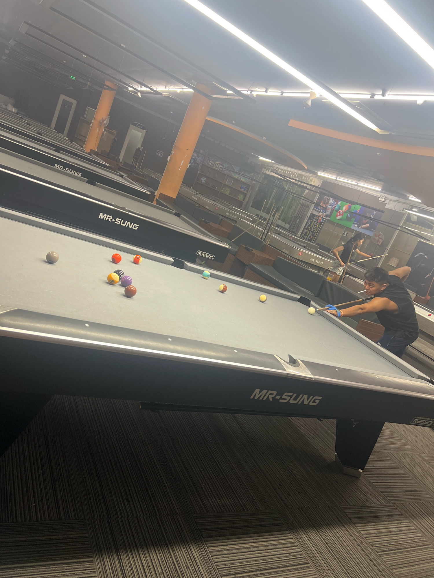 The image shows an interior view of a dimly lit pool hall, likely located in Phú Quốc, Vietnam, as indicated by the contextual information. Several pool tables, branded "MR-SUNG," are visible, with a few balls scattered on one of the tables in the foreground. A man, appearing to be of Asian descent and wearing a black tank top and dark shorts, is actively playing pool. He is positioned at the table in the foreground, holding a pool cue and wearing blue protective gloves on his left hand. His gaze is directed towards the cue ball. In the background, another person, a woman with dark hair and wearing a black t-shirt and shorts, is also visible playing pool at a table further back in the establishment. The environment is characterized by the presence of overhead lighting fixtures, orange-colored support beams, and dark-patterned carpeting on the floor.