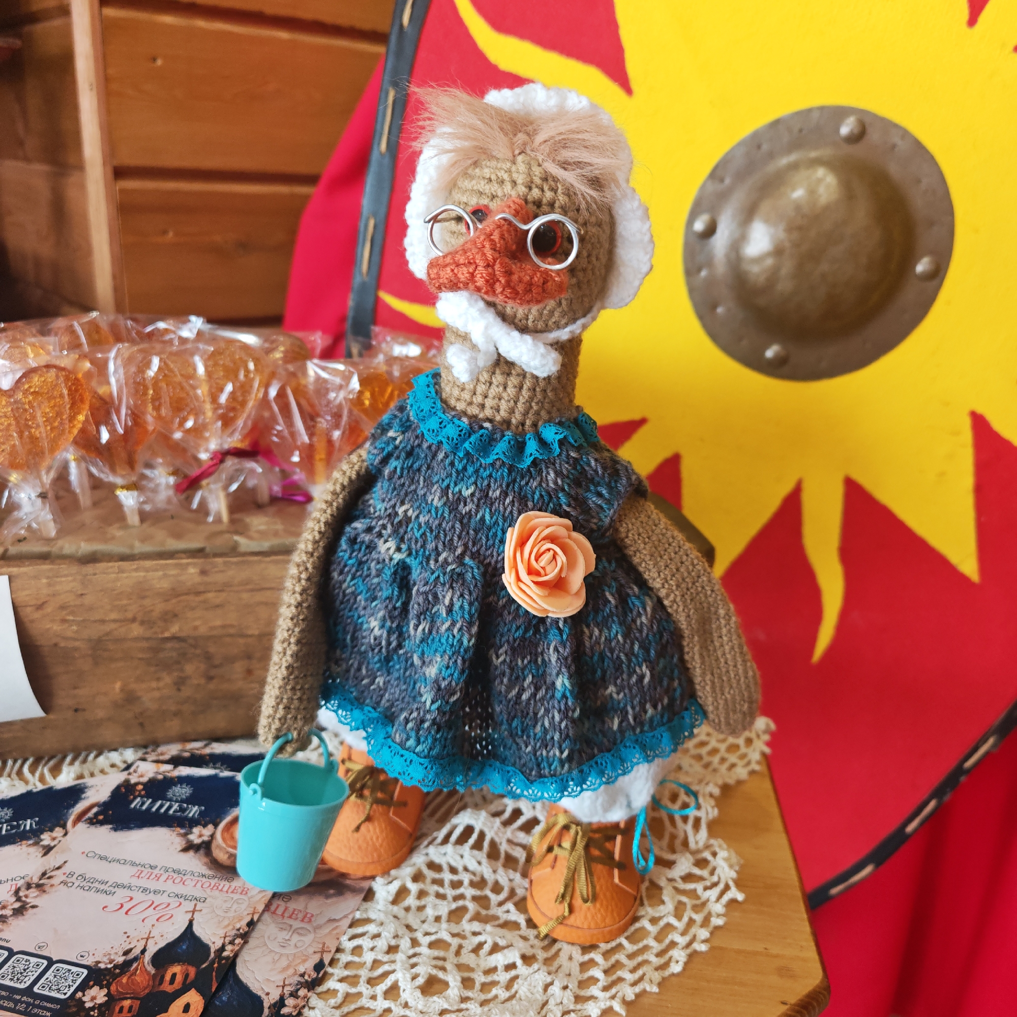 A knitted doll resembling a goose is wearing a blue knitted dress with a peach-colored rose, orange shoes with blue laces, and wire-rimmed glasses. The doll is holding a small blue bucket. The doll is sitting on a doily, along with what appear to be promotional cards. A wooden box containing individually wrapped lollipops sits behind the doll to the left. Behind the doll is a decorative shield, red and yellow in color, leaning against a wooden wall.
The promotional cards reference the city of Rostov, Russia. They advertise a special offer for residents, including a weekday discount on drinks, at a place called "Kitezh," which appears to be located at a specific address (1/2, 1 floor) not fully visible. The text on the cards is in Russian.