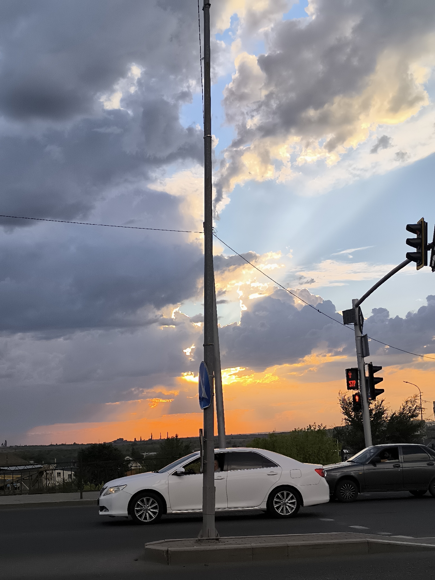 A traffic light displays a red signal for pedestrians and vehicles at an intersection in Qarağandı, Kazakhstan, during a sunset. Dark, dramatic clouds dominate the sky, with rays of sunlight breaking through, illuminating the scene with warm, golden hues. A white sedan and a dark sedan are stopped at the intersection. A tall utility pole stands prominently in the foreground, casting a shadow. In the background, a low-rise city landscape with buildings and trees is visible under the colorful sky.