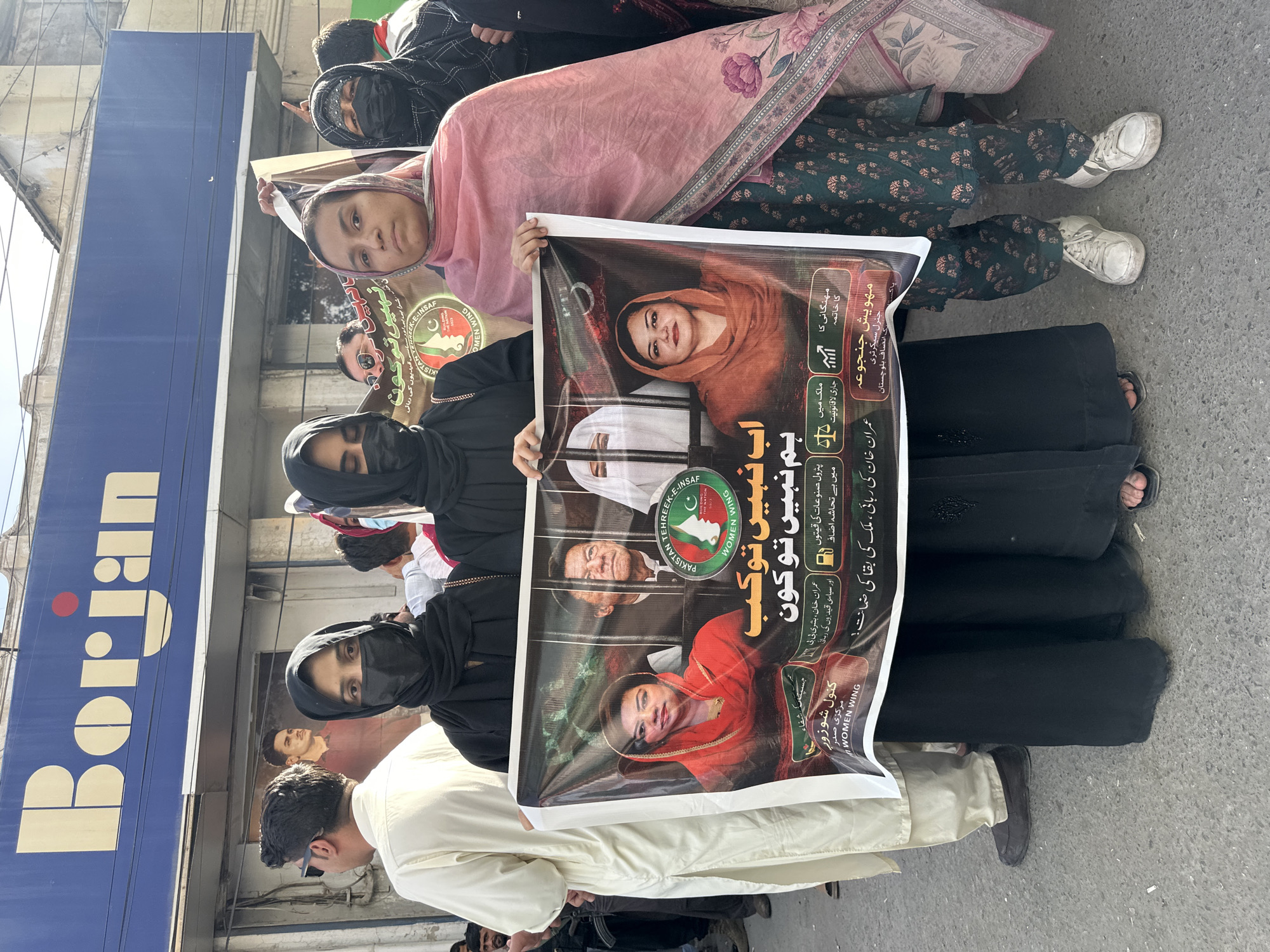 In Quetta City Tehsil, Pakistan, a group of women are protesting. The women stand outside a building with the sign "Borjan." They are holding a large poster featuring the likenesses of several people, including Imran Khan. The poster is in Urdu and mentions the political party Pakistan Tehreek-e-Insaf. One of the women wears a pink headscarf, and two are wearing black niqabs that cover their faces except for their eyes.