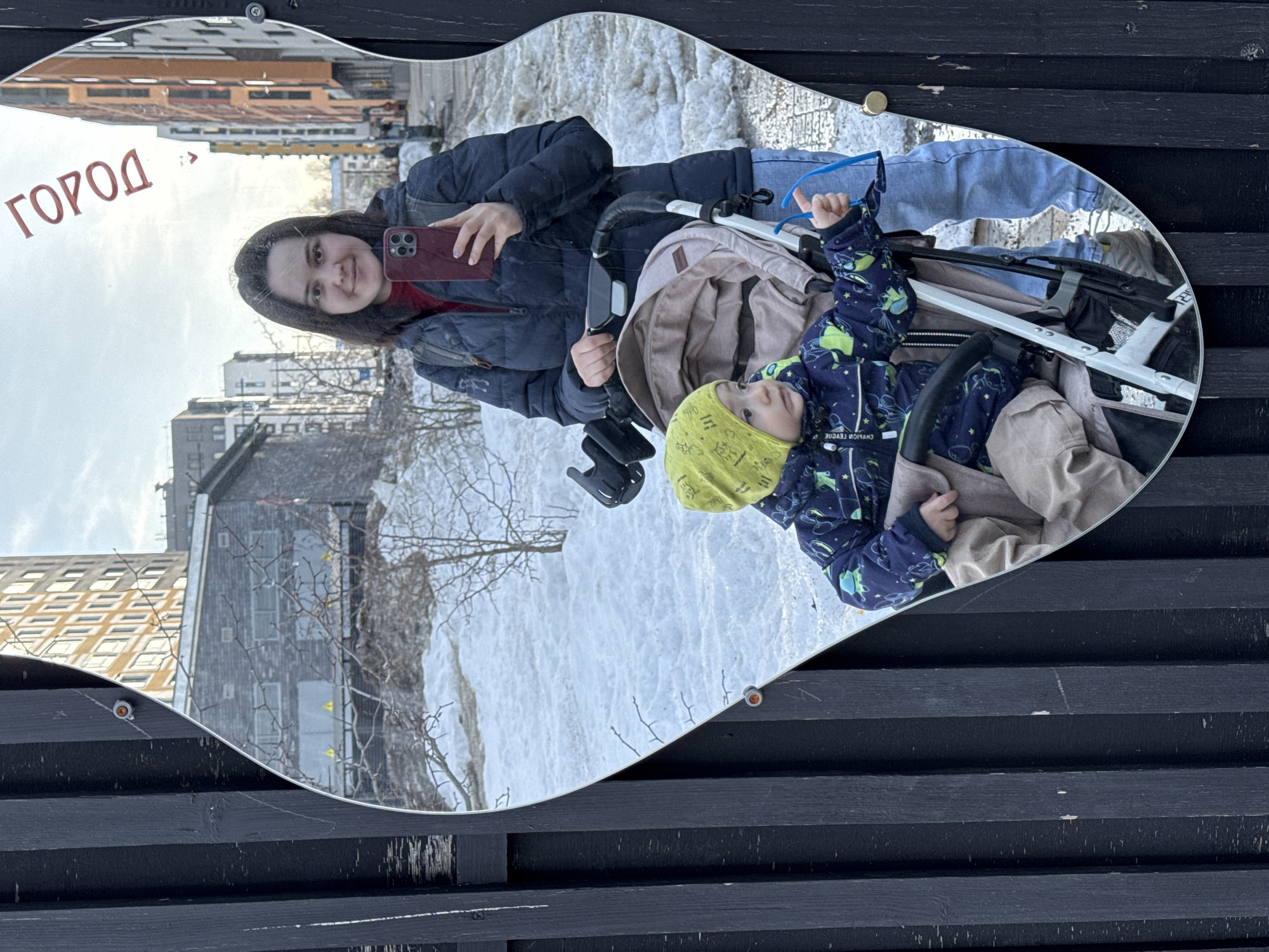 A mirror shaped like a guitar is mounted on a dark, horizontal wooden fence. The mirror reflects a woman with long dark hair taking a selfie with a red cell phone. She is wearing a dark jacket and jeans. A young child in a stroller is also reflected in the mirror. The child is wearing a yellow hat and a blue patterned jacket. The background reflected in the mirror shows a snowy scene with buildings in the city of Izhevsk, Russia. The word "город" is written in red lettering on the mirror, which translates to "city" in Russian.