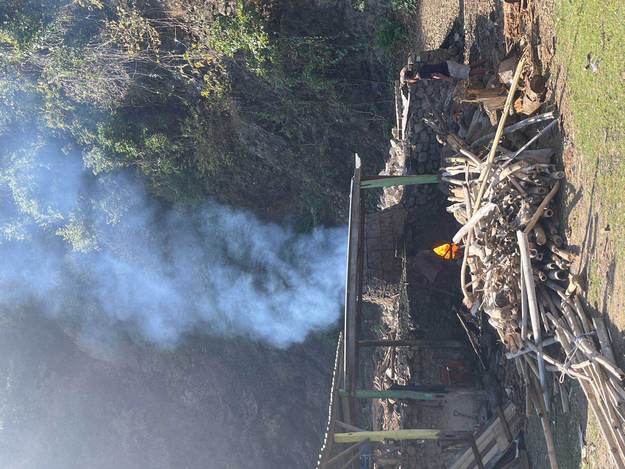 A man stands in front of a smoke-filled kiln in a rural setting in Desa Lamunga, Indonesia. The kiln is made of stone and appears to be in use, with a bright orange glow visible inside. A large pile of bamboo and wood is stacked in front of the kiln, suggesting it is being prepared for fuel. The scene is set against a backdrop of a rocky hillside covered in lush vegetation. The man is wearing a black t-shirt and shorts, and he is looking towards the kiln. The smoke billows upwards and obscures the trees and hillside behind the kiln. The overall impression is one of traditional craft and industry in a natural environment.