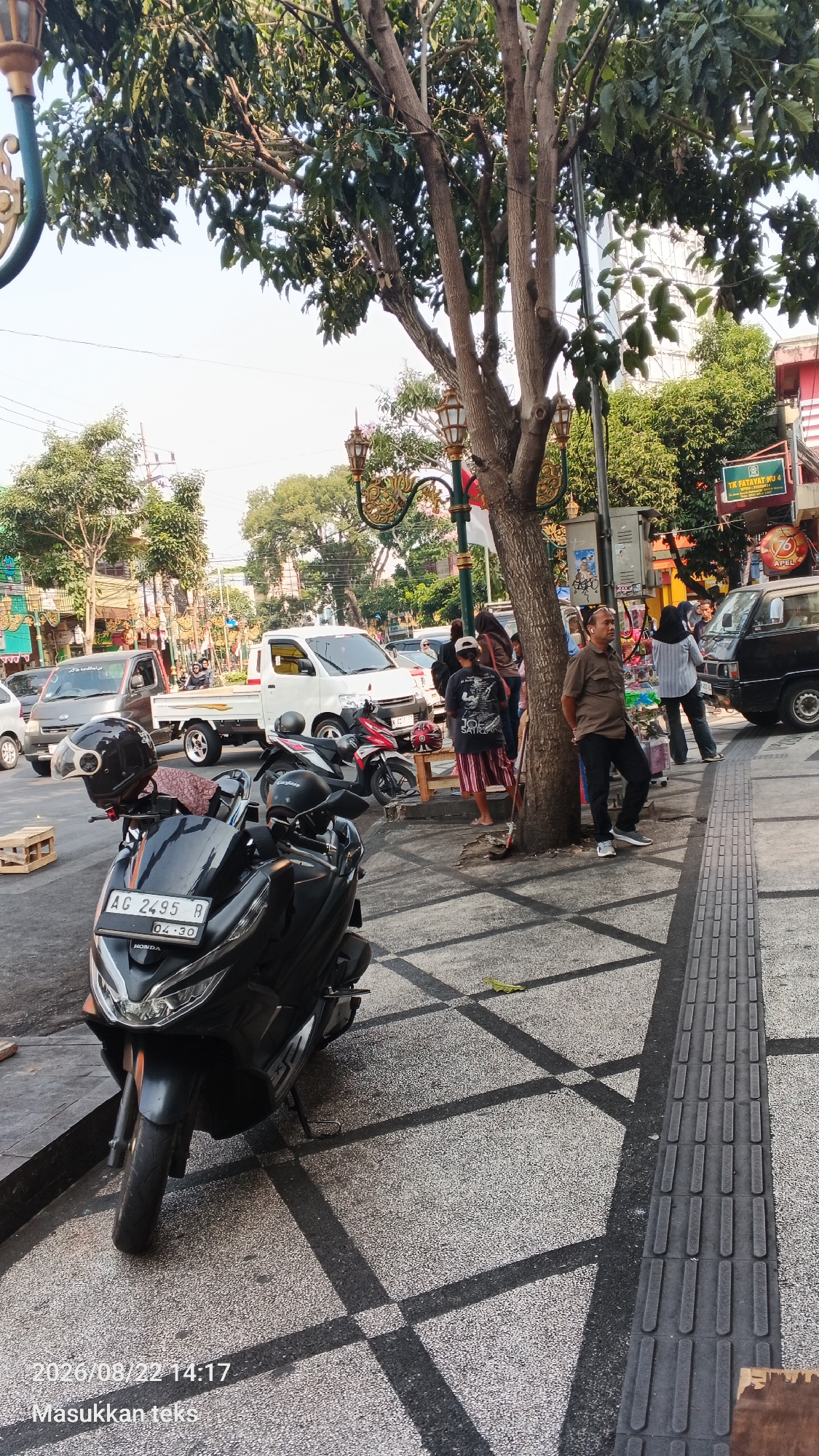 The image captures a street scene in Malang, Indonesia, with a focus on the sidewalk area in front of some buildings. In the foreground, a black Honda motorcycle is parked, with its license plate visible. To the left of the motorcycle, a wooden crate is placed on the ground. The sidewalk is paved with a speckled material and features a tactile paving strip along the edge.
Several people are present on the sidewalk and near the street. Two men are standing near a tree, one wearing a brown shirt and dark pants, the other partially obscured. A group of people, including women in headscarves, are gathered near a small table and wooden planks. Two motorcycles are parked further back on the sidewalk, one red and white, the other dark.
In the background, several vehicles are on the street, including a white pickup truck, a dark van, and a grey pickup truck. Buildings with visible signage line the street, suggesting a commercial area. A tall tree with dense foliage dominates the mid-ground, casting shadows on the sidewalk. Decorative lampposts are also visible along the sidewalk. The overall scene suggests a busy urban environment on a sunny day.