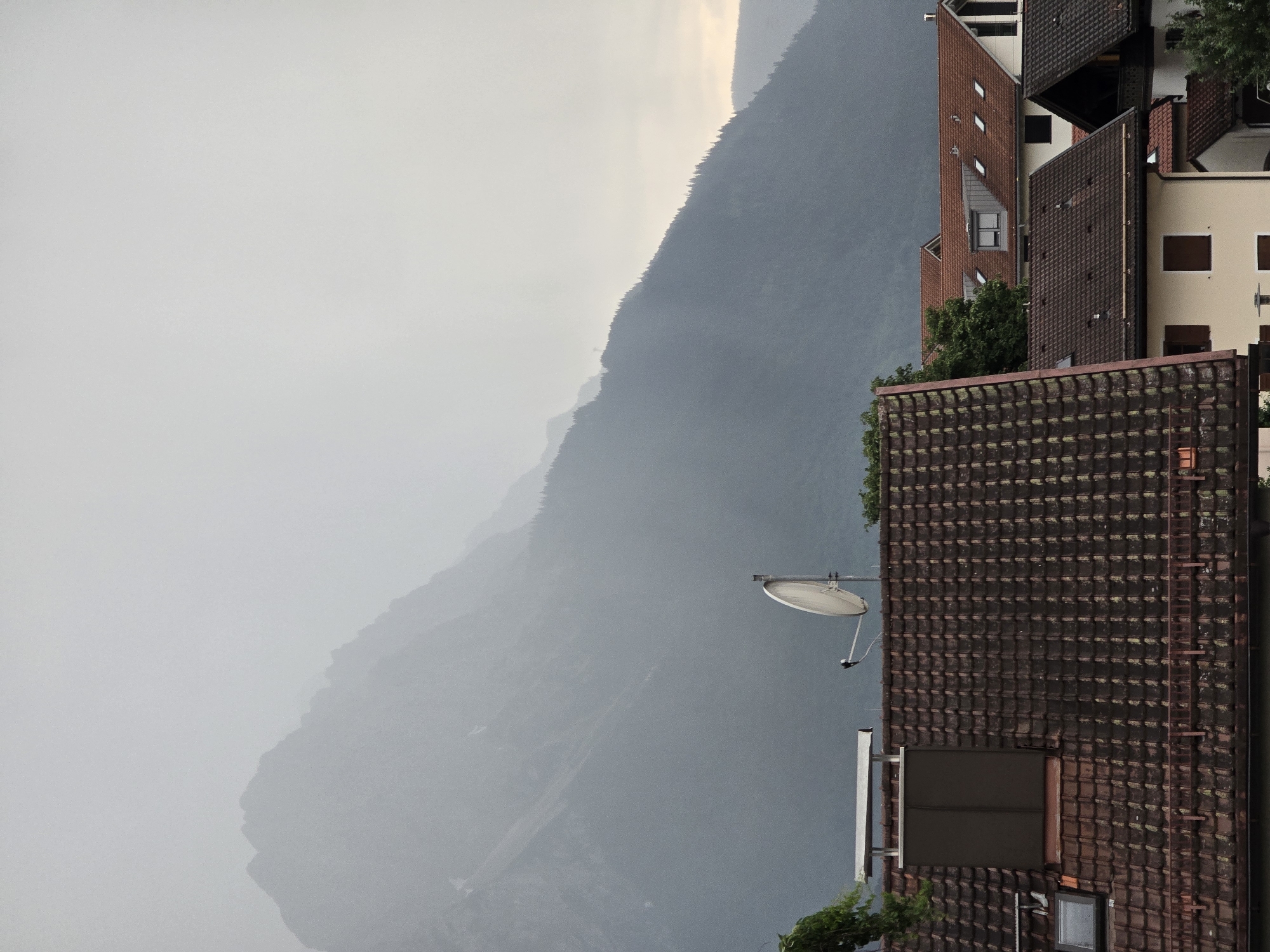The image depicts a view of rooftops in the foreground, with a large, misty mountain range in the background. The sky is overcast and a light gray. The rooftops are predominantly red-tiled, with some houses featuring dormer windows and chimneys. A satellite dish is visible on one of the rooftops. The mountain range is covered in dark green trees on its lower slopes, with the peaks shrouded in mist. The scene suggests a cloudy or foggy day in a mountainous region, possibly Garmisch-Partenkirchen, Germany, given the typical scenery of the Bavarian Alps. No people are visible in the image.