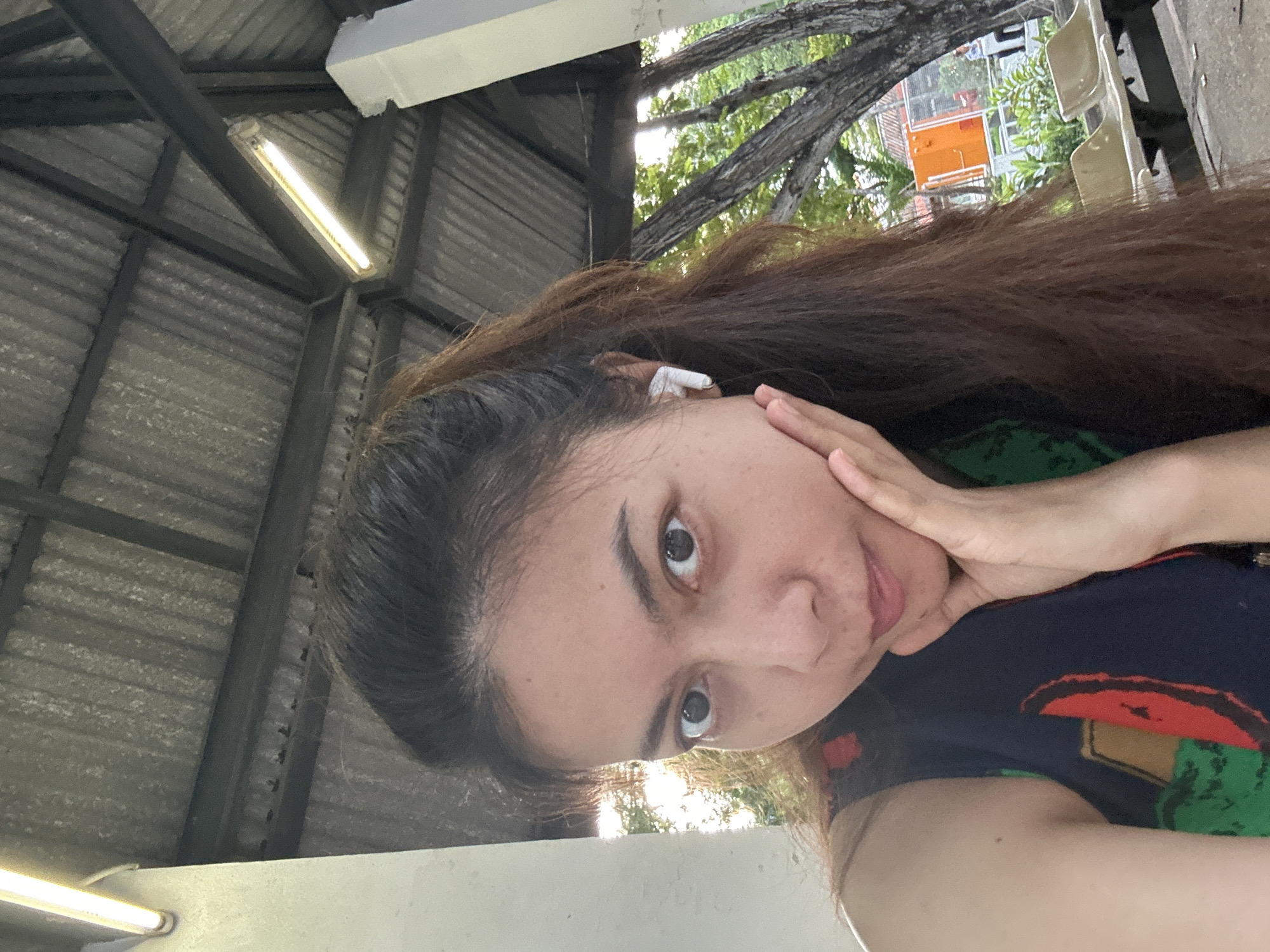 A woman with long, dark brown hair pulled into a ponytail, wearing a dark blue sleeveless top with a colorful pattern, is taking a selfie. She is resting her right hand on her cheek. She is wearing a white wireless earbud in her right ear. The background appears to be an outdoor covered area with a corrugated metal roof supported by beams and pillars. Fluorescent lights are visible on the ceiling. Outside the covered area, trees and some outdoor seating are visible. The scene suggests a public area like a bus stop or waiting area, possibly in Tha Khlong Town Municipality, Thailand, given the vegetation and architecture typical of the region.