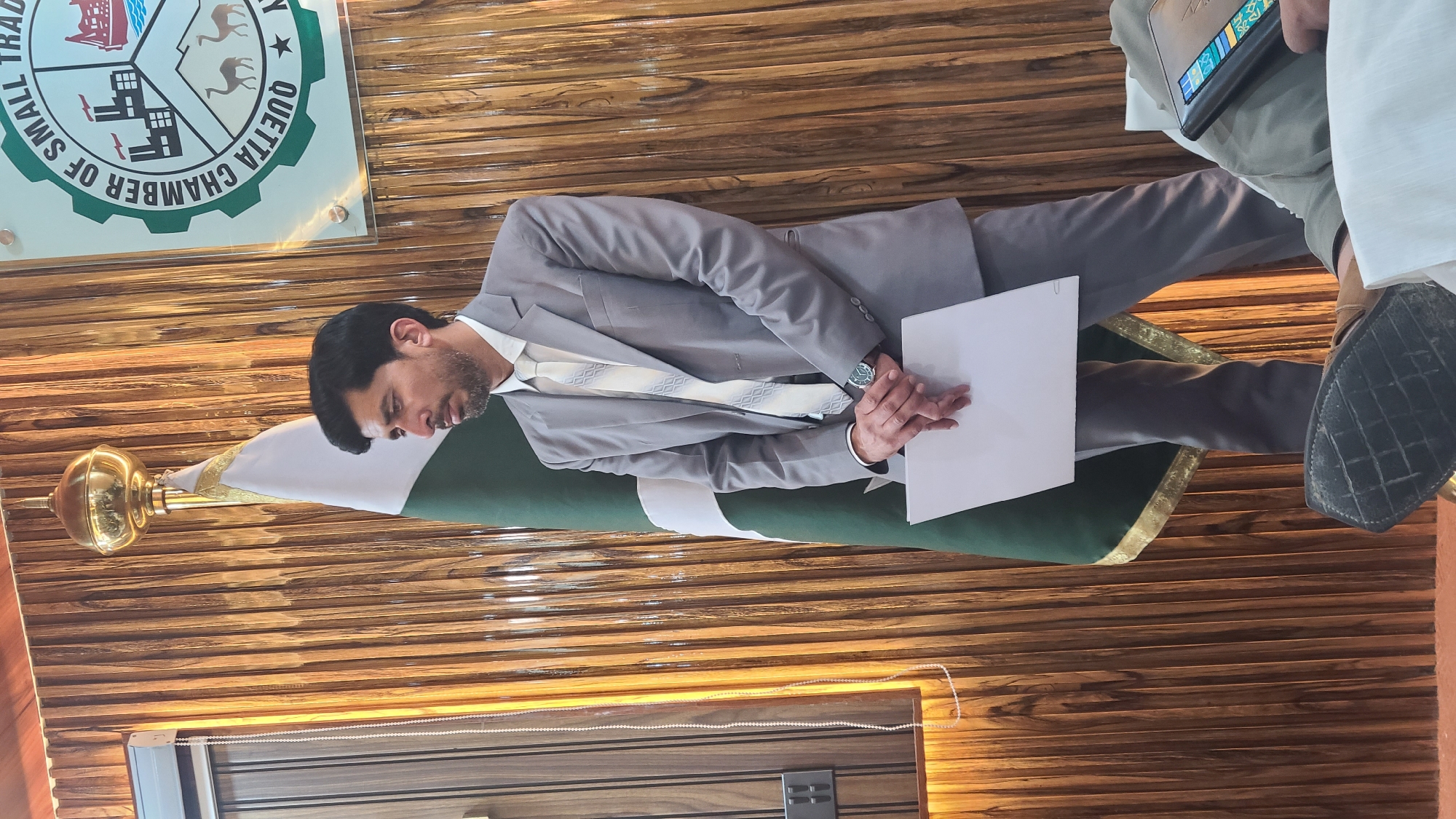 A man in a grey suit is standing in front of a flag of Pakistan. He is holding a white paper. The room has wood paneling and there is a sign for the Quetta Chamber of Small Trade. This suggests the location is in Quetta City Tehsil, Pakistan, as Quetta is a city within that tehsil.