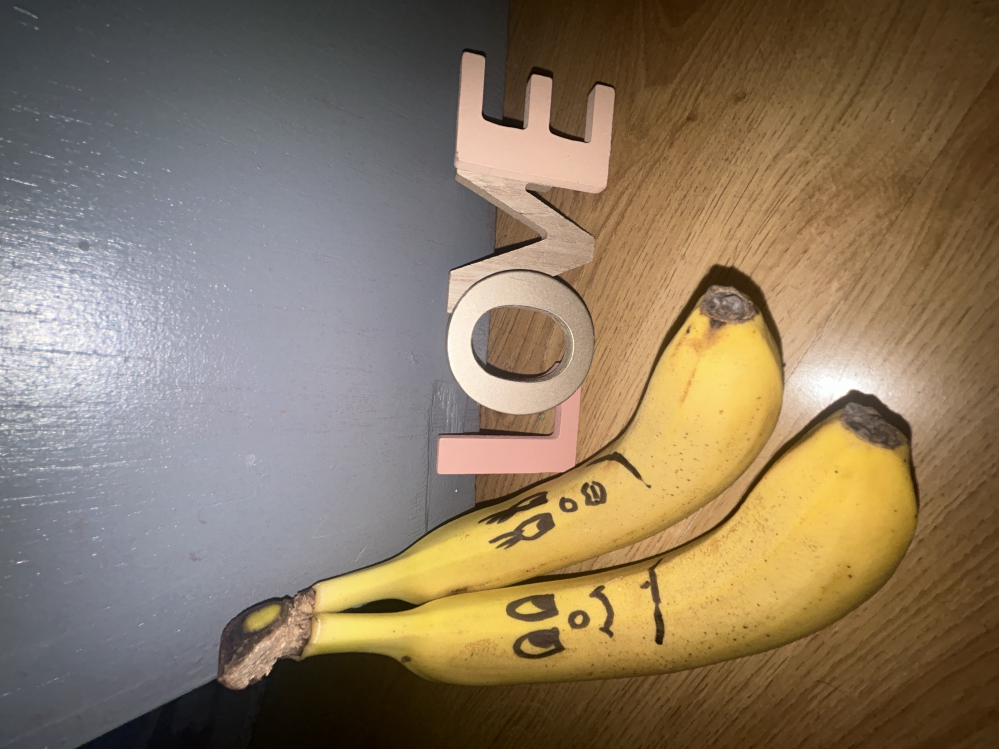 The image shows two bananas with faces drawn on them, lying next to the word "LOVE" constructed from wooden letters. The letters are stacked vertically with "E" on top, then "V", "O", and "L" on the bottom. The background is split between a gray surface on the left and a wooden surface on the right.
