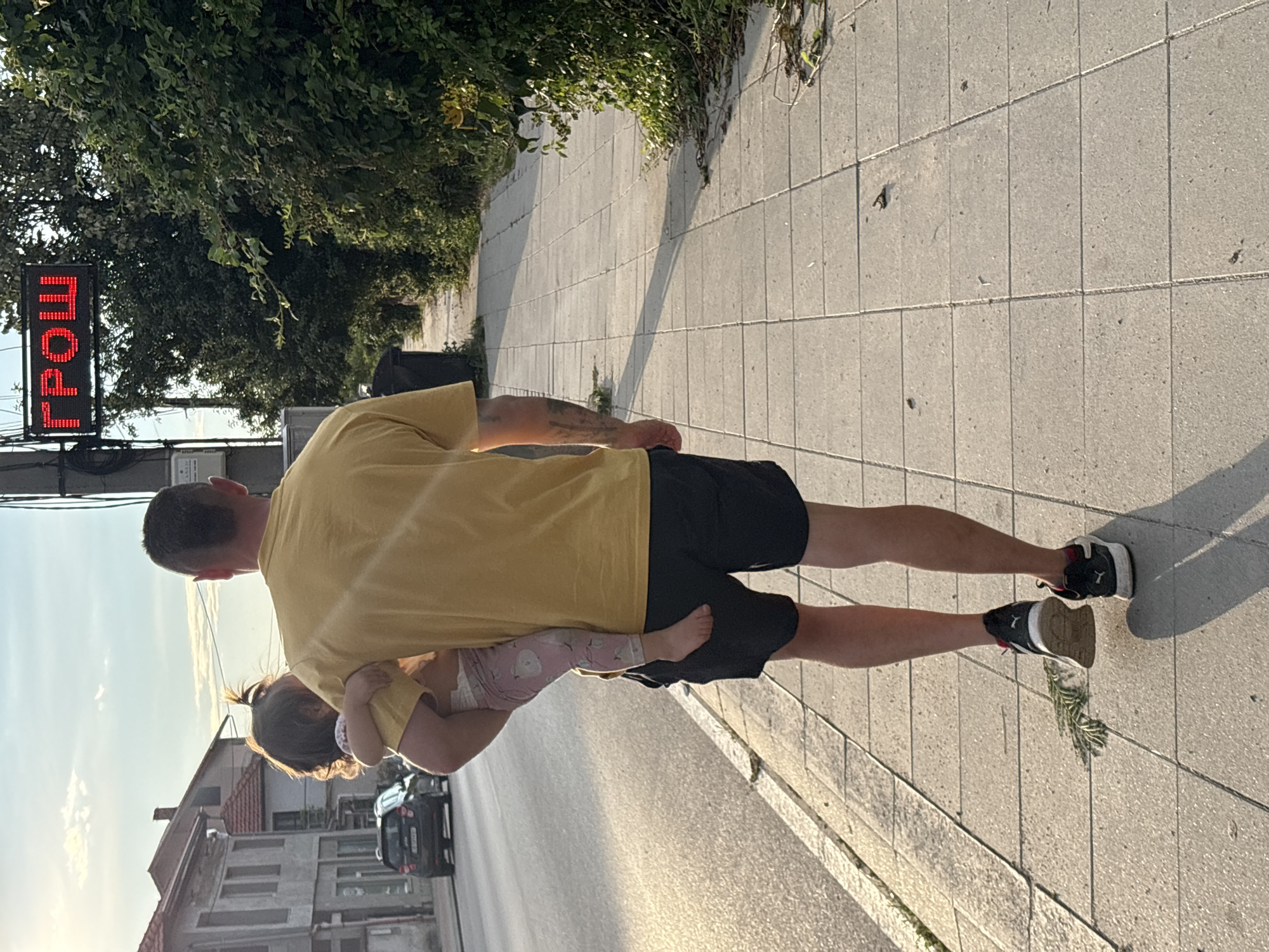 A man wearing a yellow t-shirt and black shorts carries a young child in his left arm. The man is walking on a sidewalk next to a street. The child is facing away from the camera and is wearing a pink outfit. In the background, there are buildings, trees, and a digital sign that displays red letters. The image appears to be taken during the daytime, possibly in the late afternoon, given the lighting. The sign in the background displays the word "ГРОШ" in Cyrillic, which translates to "GROSH" in English. This is likely a street name or a business name in Oreshak, Bulgaria. The sidewalk is made of concrete tiles, and there is some debris on the ground. A dark-colored car is visible on the street in the distance.