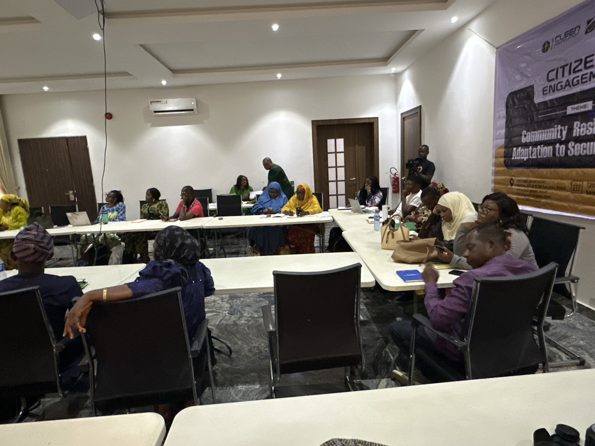 A conference room in an unknown city features attendees seated around rectangular tables, engaged in a meeting or workshop. A banner at the side of the room indicates the theme "Citizen Engagement: Community Resilience Adaptation to Security." The banner also lists an address that indicates the city of the event is Unknown. Participants are diverse in age, ethnicity, and attire, with some wearing traditional garments and others in business casual attire. The room contains multiple doors and a fire extinguisher mounted on the wall. The lighting is bright. The floor has a grey marble design.