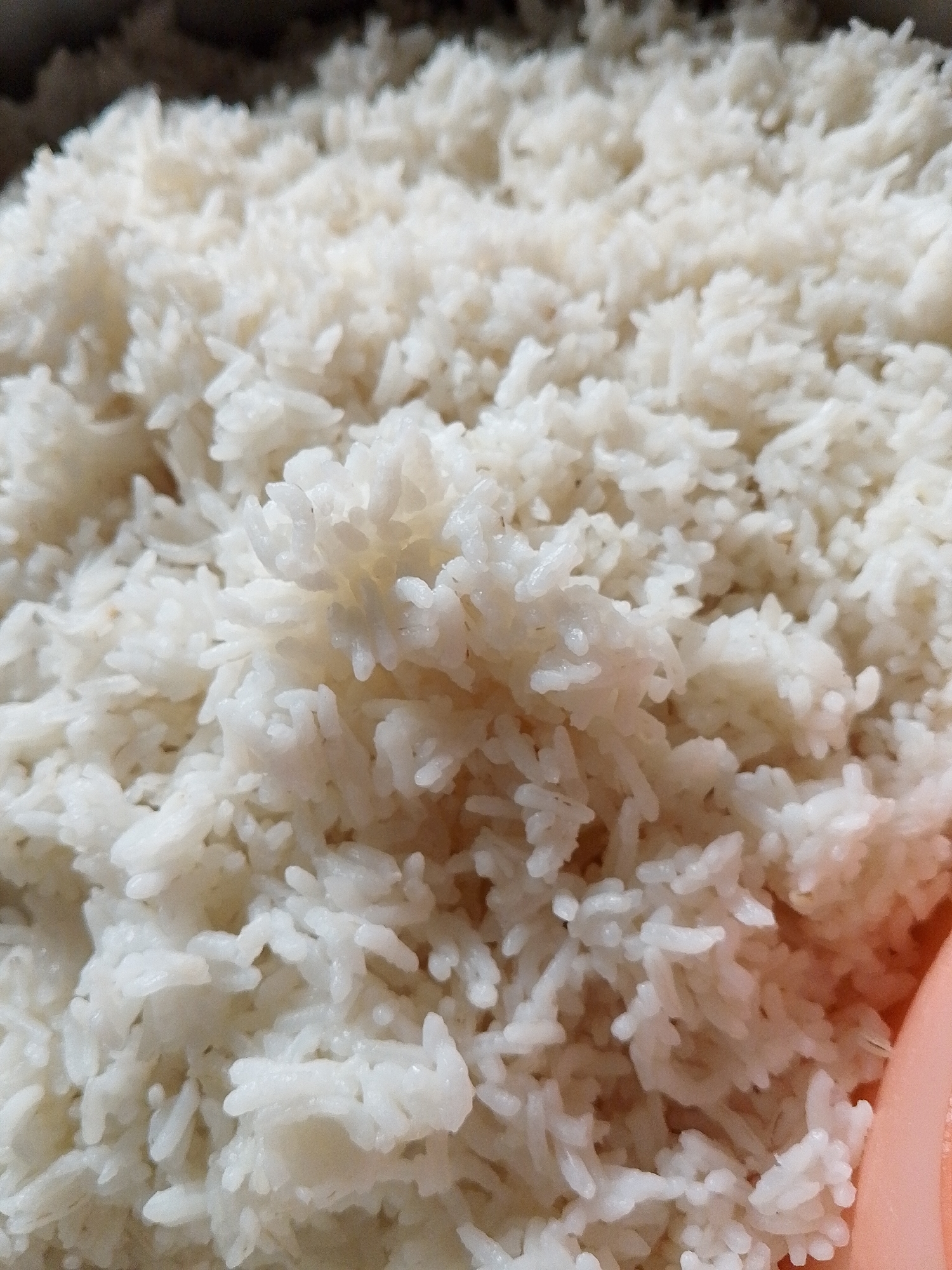 A close-up, overhead shot shows a large quantity of white, cooked rice filling most of the frame. The rice is piled high, with individual grains visible, and appears to be moist and fluffy. A sliver of a pink object, possibly a bowl or a utensil, is visible on the right edge of the image. The background is dark and out of focus, suggesting the rice is in a pot or container. The image provides no information about Lhokseumawe, Indonesia.