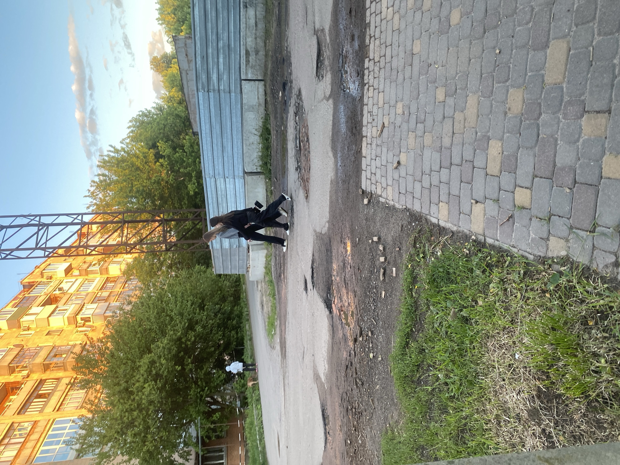 The image depicts an urban scene in Poltava, Ukraine. A woman is walking on a paved road, which is in disrepair with potholes, toward a second person walking a dog. A section of the walkway is paved with stones. Adjacent to the road is green grass and trees. A large, multi-story building is visible in the background, alongside a metal structure and corrugated metal fencing. The sky is partially visible with some clouds.