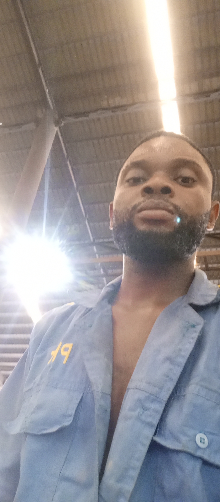 A close-up photo shows a dark-skinned man with a beard wearing an unbuttoned light blue work shirt with yellow lettering on the pocket. The location appears to be indoors, possibly a warehouse or industrial space, judging by the corrugated metal ceiling and steel support beams visible in the background. There are bright sources of light near the ceiling. The man is looking directly at the camera. The location in Magboro, Nigeria, cannot be confirmed from the visual information.