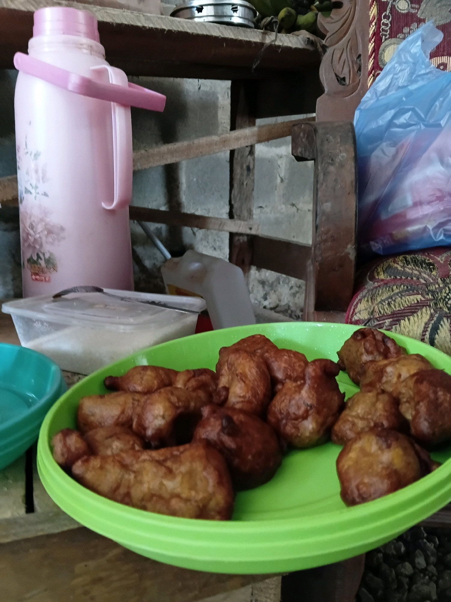 A pink plastic thermos with a light pink handle is visible on the left side of the image. In front of the thermos is a clear plastic container with a spoon on top, filled with a white granular substance, likely sugar. A bright green plastic platter filled with fried dough pieces, possibly pisang goreng (fried bananas), is placed in the foreground. To the left of the platter, a teal plastic bowl is partially visible. The background consists of a rustic, outdoor setting with a brick wall, wooden planks, and a blue plastic bag. The overall impression is a casual, domestic scene, likely in a rural or semi-rural area. The presence of pisang goreng suggests a traditional Indonesian setting.