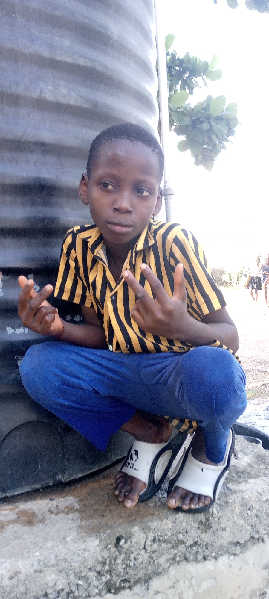 In Adankolo, Nigeria, a young dark-skinned boy wearing a yellow and black striped shirt and blue pants is crouched down. He is wearing white and black Adidas sandals. He is making hand gestures, with his fingers extended in a "peace" sign. He is leaning against a large, black tank. The background is partially obscured by bright light but includes greenery and two distant figures of people walking. The ground is cracked and stained.